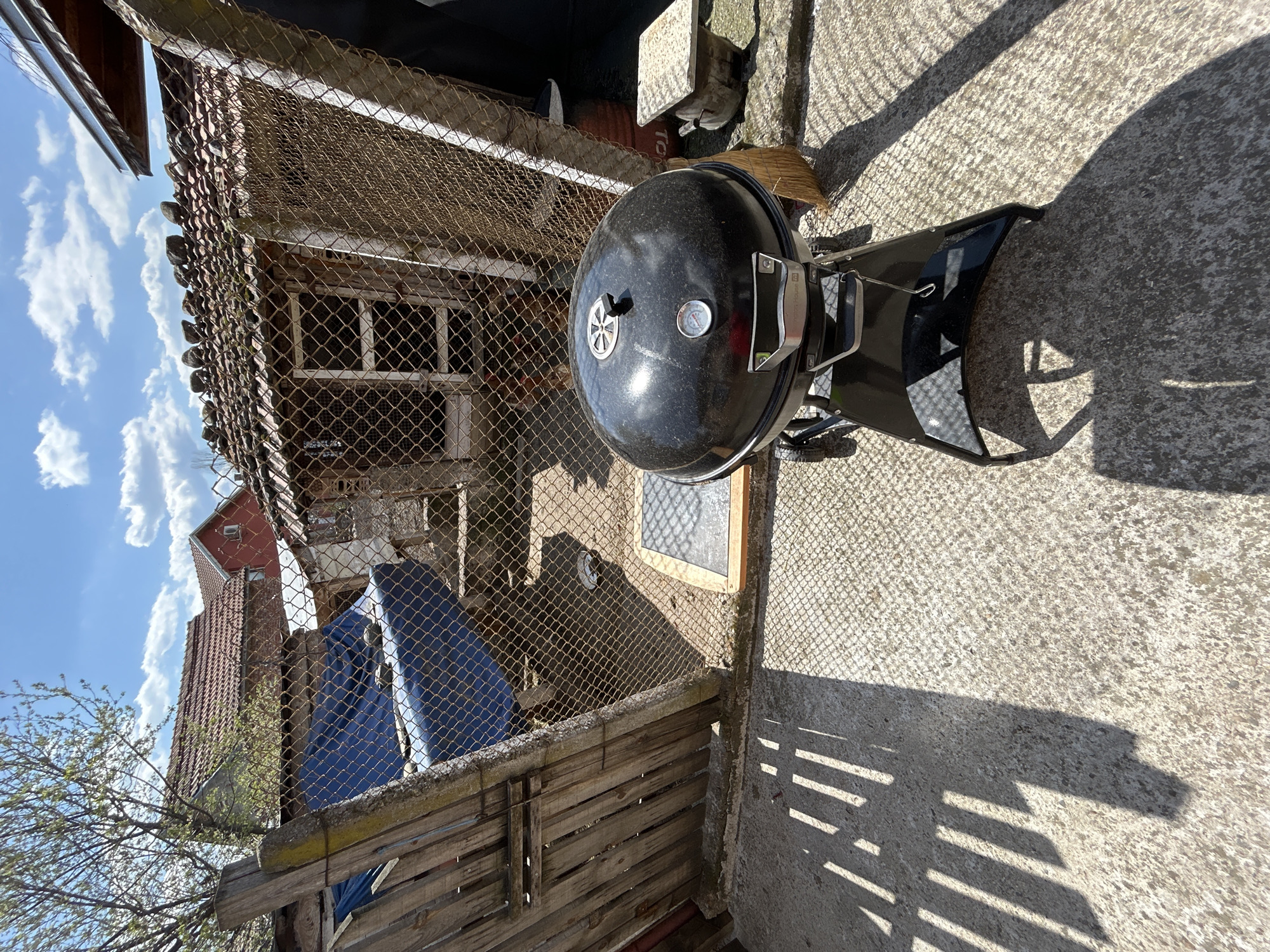 The image shows a backyard scene in Gabarevo, Bulgaria. A black charcoal grill is positioned on a concrete surface next to a dilapidated wooden fence topped with chain-link fencing. Behind the fence, there's an old, weathered shed-like structure with a tile roof and a fenced-in window. A blue tarp is partially visible behind the fence. In the background, sections of neighboring buildings with red tile roofs can be seen under a blue sky with scattered clouds. The sun is shining, casting shadows from the fence and the grill onto the concrete.
