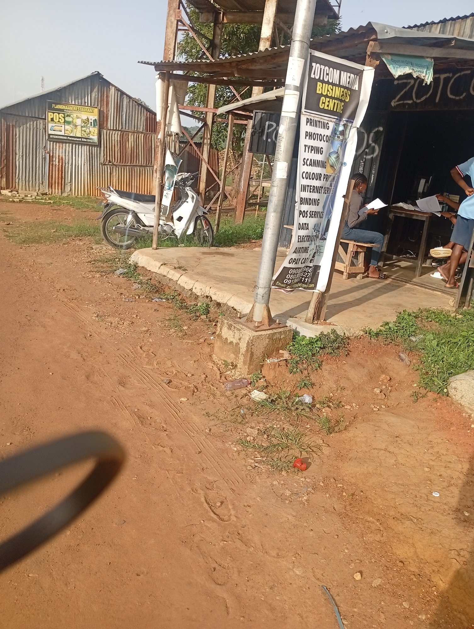 The image depicts an outdoor scene in a rural or semi-urban setting. In the foreground, a dirt path with tire tracks is visible, leading towards a concrete curb and overgrown vegetation. A motorcycle is parked near a corrugated metal building. To the right, a business called "ZOTCOM MEDIA BUSINESS CENTRE" is advertised on a banner attached to a pole. Inside the establishment, two individuals are seen at a desk with papers. One person is sitting and looking at documents, while another appears to be standing. The overall impression is of a small commercial operation situated in a somewhat dilapidated environment. The sky is clear and bright.