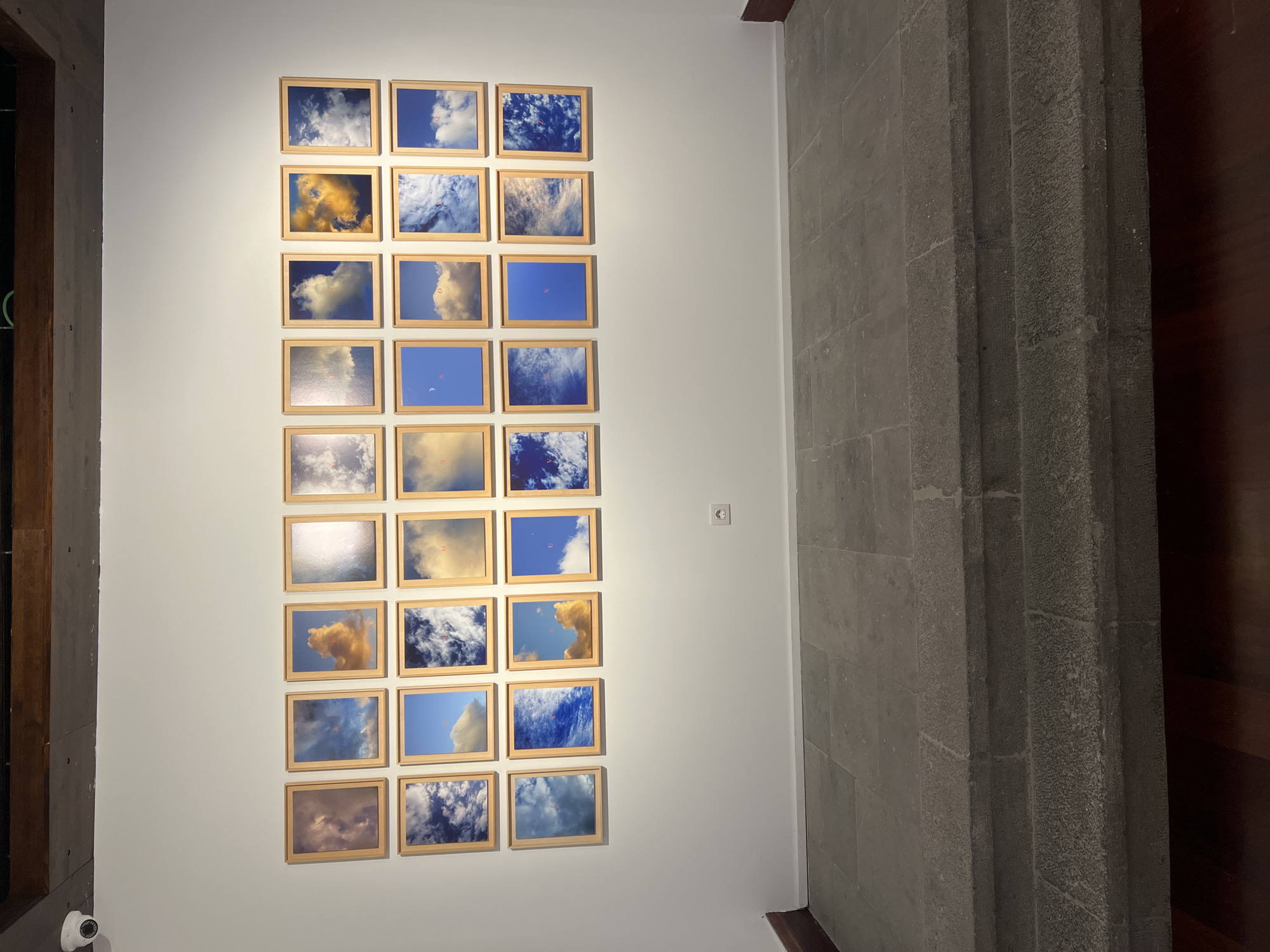 The image presents an interior view, likely an art gallery or exhibition space. A collection of twenty-four framed pictures is mounted on a white wall. The pictures depict scenes of clouds and skies, with the frames appearing to be made of light-colored wood. The pictures are arranged in a grid pattern on the wall. The overall aesthetic suggests a contemporary art installation.
To the right of the white wall is a section of gray stone or concrete wall with a textured surface. Further to the right, there is a wooden panel, possibly part of a door or another architectural element. A small white electrical outlet is visible on the white wall, near the stone wall. On the left side of the image, part of a dark-colored frame or wall is visible. A small security camera is mounted on the wall at the bottom left corner.
Based on the architectural style, materials (stone and wood), and the presence of contemporary art, it is plausible that this scene is located in Las Palmas, Spain, but without specific architectural landmarks, signage, or distinctive cultural elements, a precise location confirmation within Las Palmas is not possible.