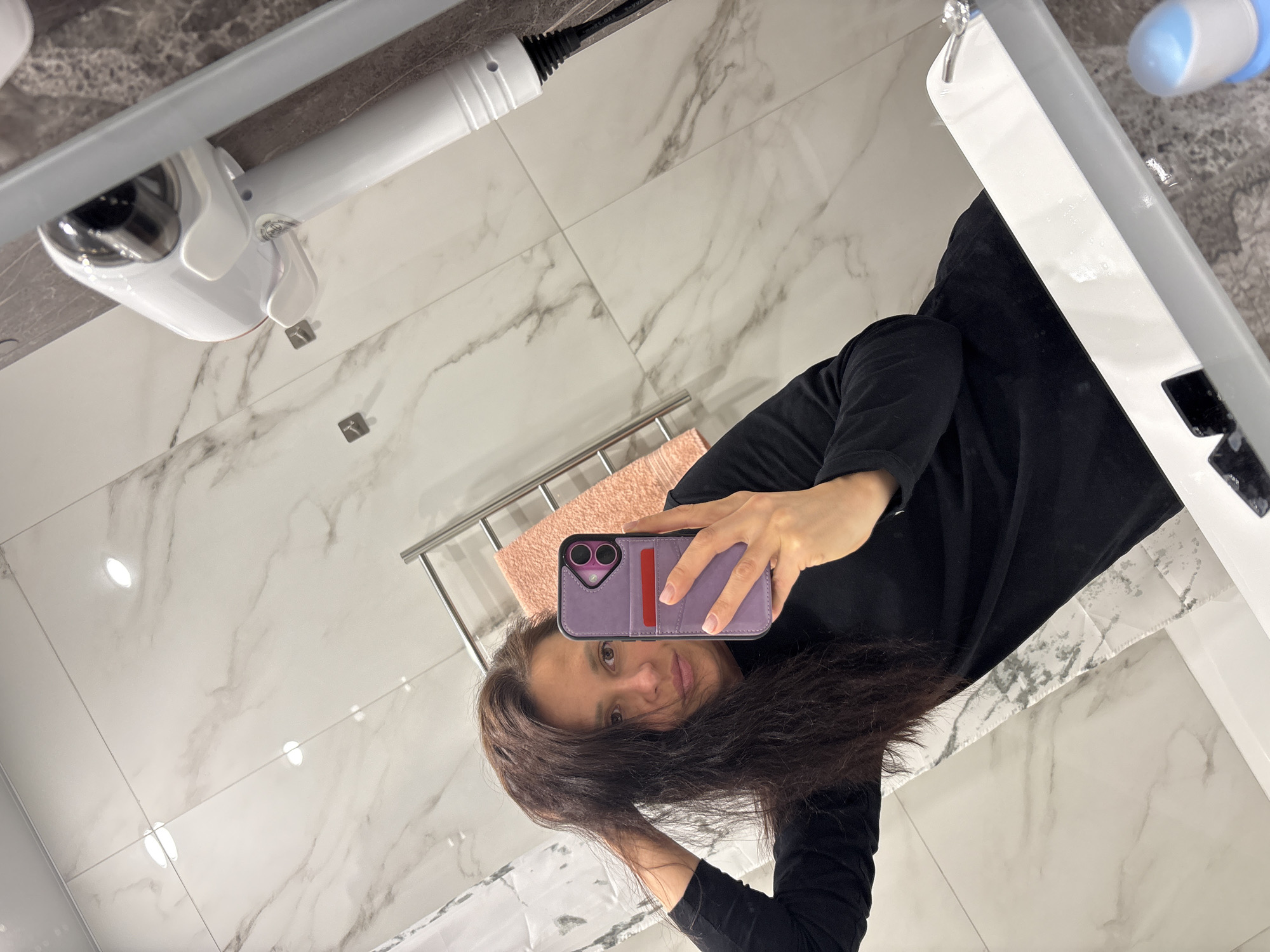 This mirror selfie depicts a woman in a modern bathroom in Kazan, Russia. The woman, who has long, dark brown hair and a neutral expression, is wearing a black long-sleeved top. She is holding a smartphone in a purple case with a red accent, using it to take the picture. Her hair appears slightly disheveled, suggesting she might be in the process of styling it or has recently finished showering.
The bathroom features bright, white walls adorned with grey marble-patterned tiles, giving it a clean and contemporary look. Visible elements include a white sink or vanity unit with a silver faucet. Above the sink, a white hairdryer or hair styling appliance is mounted on the wall. A chrome towel rack holds a folded pink or peach-colored towel, and small, square wall hooks are also present. On the right side of the sink, a blue and white object, possibly a hygiene product or electric toothbrush, is visible. The lighting in the room is artificial, typical for an indoor bathroom, making it impossible to determine the time of day. No legible text is visible in the image.