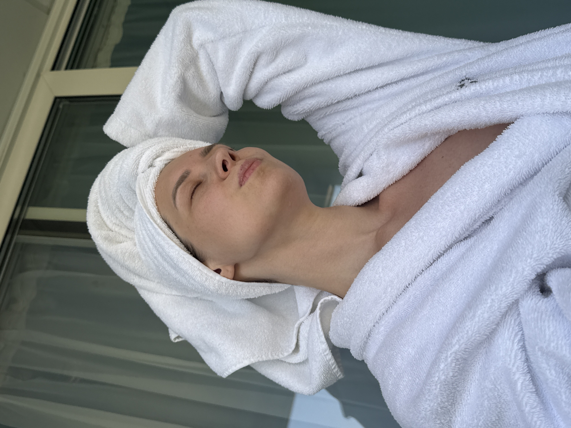 In Nebug, Russia, a young woman with her hair wrapped in a white towel and wearing a white bathrobe is photographed indoors. She is leaning back with her eyes closed and her mouth puckered, appearing relaxed and perhaps enjoying a moment of self-care. Her right arm is raised, with her hand resting on the back of her head. The background suggests an indoor environment with windows and sheer curtains, hinting at a hotel room or similar accommodation. The lighting is soft, possibly natural light filtering through the windows.