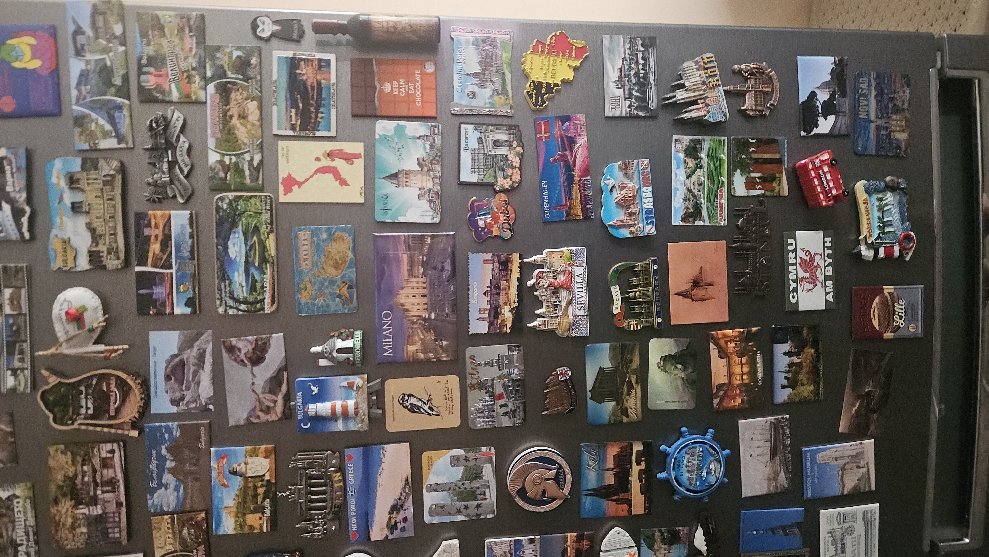The image shows a collection of magnets affixed to a refrigerator door. The magnets appear to be souvenirs from various locations, including cities, countries, and specific landmarks. Prominent locations represented by the magnets include Milan, Istanbul, Seville, Copenhagen, Budapest, Prague, London, and Novi Sad. Additionally, magnets representing Bulgaria and Romania are visible. One magnet reads "KEEP CALM EAT CHOCOLATE." The magnets vary in size, shape, and material, some appearing to be made of plastic, metal, or ceramic. Specific location in Plovdiv is not apparent.