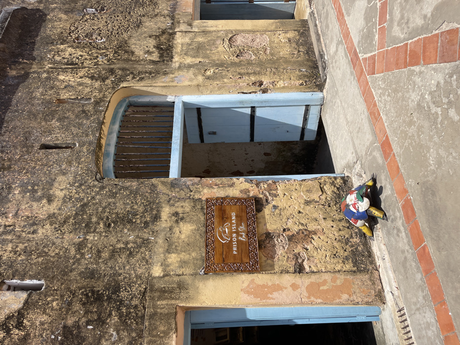 The image displays the weathered exterior of a building in Bububu, Tanzania, under bright sunlight. The primary structure features rough, discolored yellowish-brown plaster walls exhibiting signs of age and wear. Three light blue wooden doors are visible: one partially open on the far left, a central arched doorway with an open barred gate above a partially ajar double door, and a partially visible closed door on the far right. A wooden sign, horizontally oriented and framed with intricate carvings, is affixed to the wall between the left and central doors. The sign reads "PRISON ISLAND Book Shop" in white text, accompanied by a white graphic depicting a tortoise or turtle. On the paved ground beneath this sign, a multi-colored metallic sculpture of a tortoise or turtle rests. Its shell is painted in a patchwork of red, white, blue, and green, while its head and legs are metallic yellow. The ground surface consists of concrete or stone with reddish-brown rectangular tiles forming geometric patterns, and a metal drain grate is visible near the left door. No individuals are present in the scene.