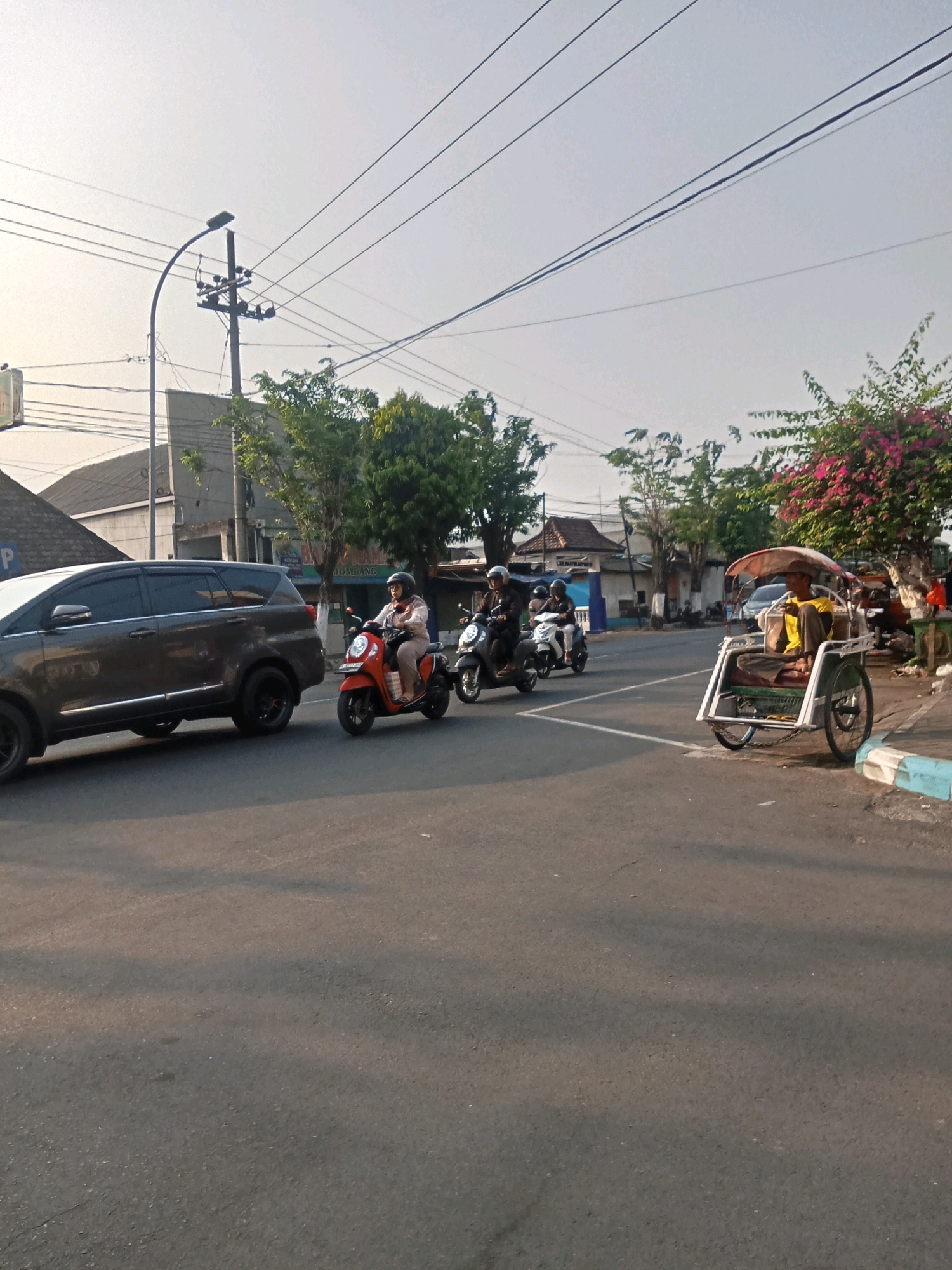 In Kaliwungu, Indonesia, a two-lane asphalt road is depicted during daylight hours, with a clear sky and soft sunlight creating shadows on the pavement. On the left, a dark gray SUV is traveling on the road. To the right of the SUV, a woman wearing a helmet and light-colored clothing rides a red scooter. Behind her, two more individuals on scooters, also wearing helmets, are visible. In the right lane, a pedicab with a driver in a yellow shirt and brown pants is stationary. Buildings with various roofs and some vegetation line the right side of the road, including a tree with pink flowers. Overhead, utility poles and power lines stretch across the sky.