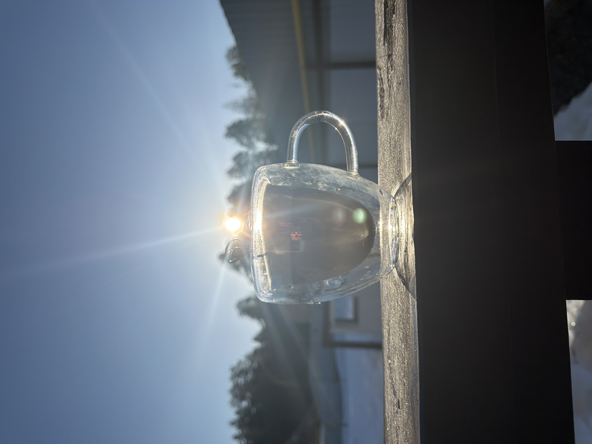 A clear glass mug with a handle sits on a weathered wooden surface under a bright sun, producing strong lens flare and reflections. The mug contains liquid. Behind the mug, blurred greenery and building structures are visible against a blue sky. The photograph appears to have been taken outdoors, likely in Chaikovsky, Russia.