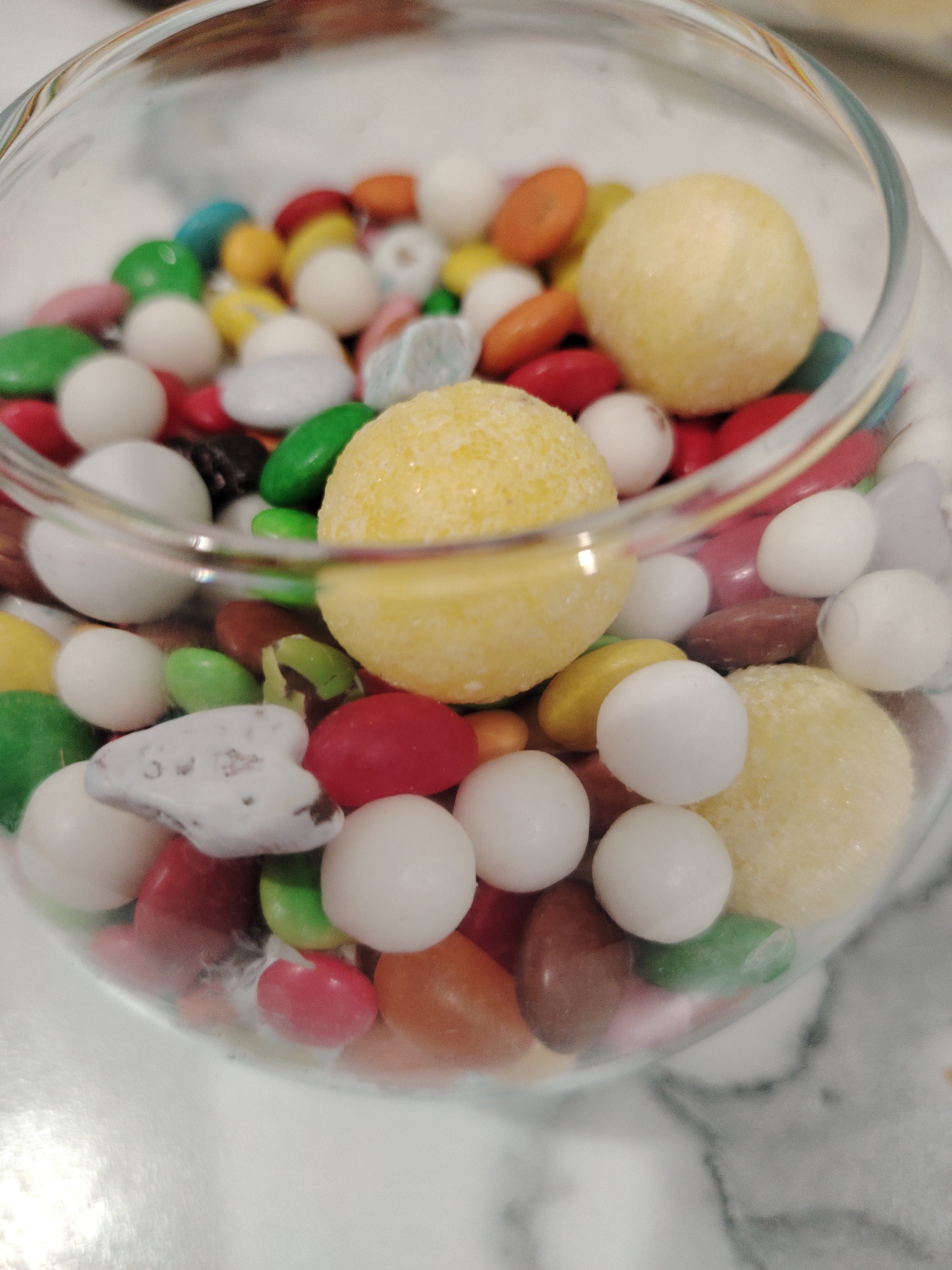 The image shows a glass bowl filled with a variety of candies. There are small, oval-shaped candies in multiple colors including red, orange, green, white, and brown. There are also larger, round, white candies, and even larger, round, light yellow candies with a textured surface. The bowl appears to be sitting on a white or light-colored surface.
There is no verifiable information that this image was taken in Sarapul, Russia.