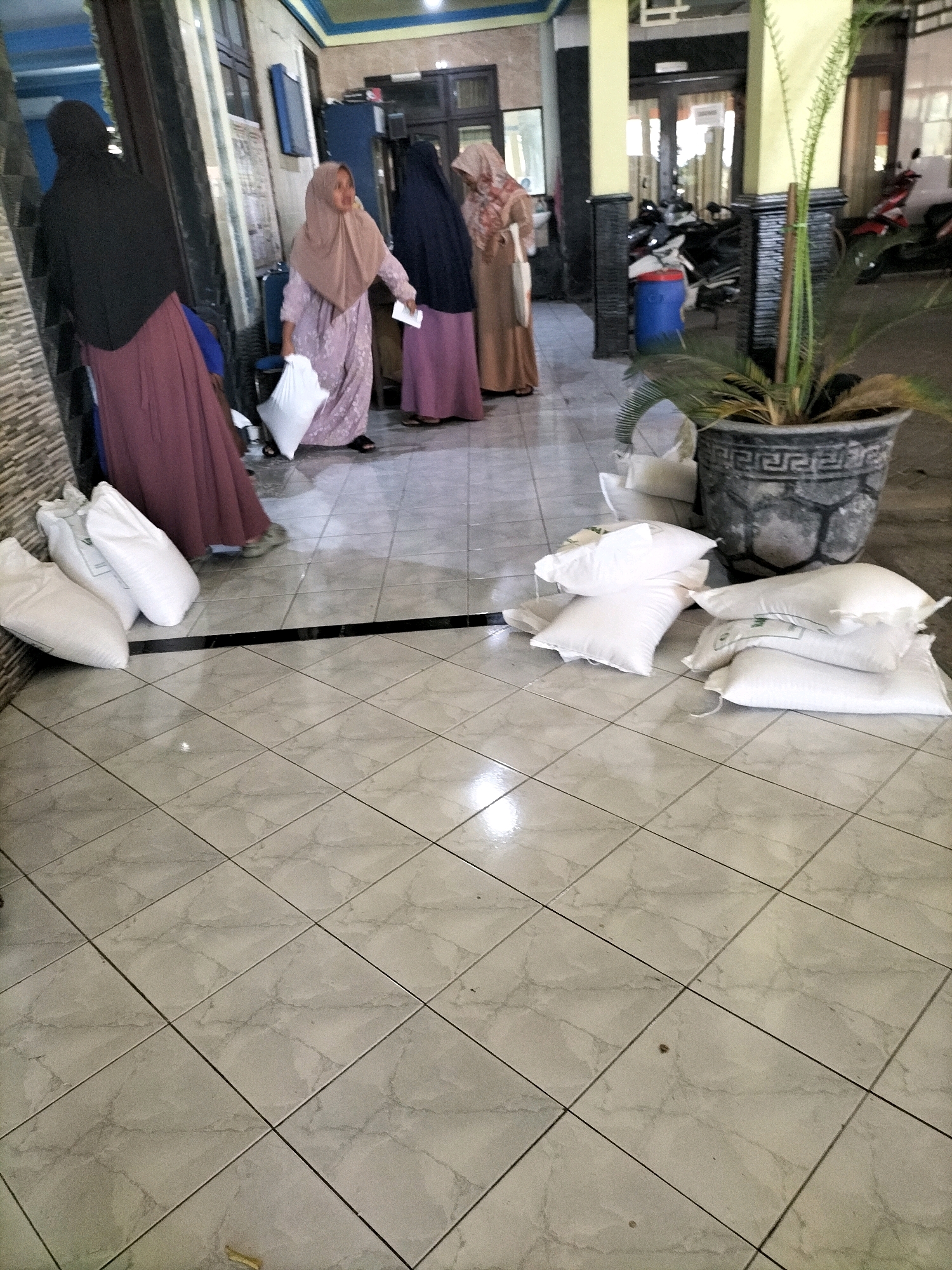 In a tiled hallway in Sembayat, Indonesia, four women are visible, three of whom are wearing headscarves. Two women are standing near a doorway on the left, one dressed in a black abaya and a maroon skirt, and the other in a light purple abaya and a light brown headscarf. The woman in the purple abaya is holding a white plastic bag. Further down the hallway, two more women are standing together. One is wearing a dark blue abaya and a light brown headscarf, and the other is wearing a light brown and orange patterned abaya and a light brown headscarf. In the foreground, several white sacks are piled on the floor. Behind the women, motorcycles are parked. A large potted plant stands near a pillar on the right side of the image.