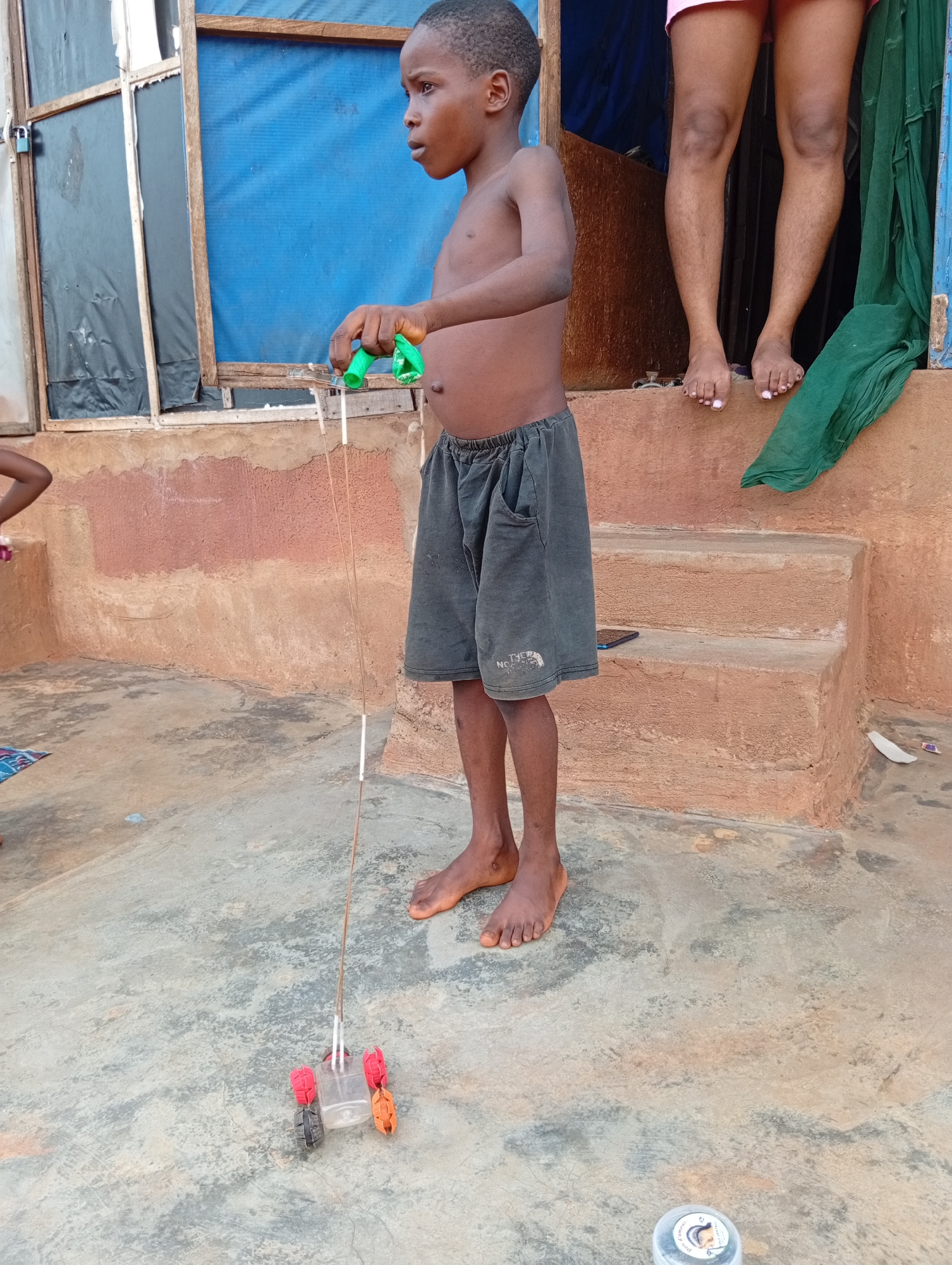A young, shirtless boy, appearing to be between five and ten years old, is depicted playing outdoors in Ogidi, Nigeria. He wears dark grey shorts, which have a partially visible "THE N FACE" logo, likely indicating "THE NORTH FACE." Barefoot, he stands on a worn concrete surface that shows cracks and discoloration.
The boy is absorbed in playing with a homemade pull-along toy car. The car is fashioned from a clear plastic container, serving as its body, with red and orange bottle caps or plastic pieces used as wheels. He guides the toy using a long, thin stick or rod attached by strings, his gaze focused downward on the car, indicating concentration.
Behind the boy is a textured, light brown/ochre-colored wall with darker patches, possibly from weathering. Two concrete steps lead up to a raised platform or doorway. On this platform, the bare legs and feet of another person, wearing pink shorts and with light-colored nail polish on their toes, are visible in the upper right. A draped green fabric hangs from a doorway or window to their right. A small portion of another person's lower body is visible on the far left.
The background includes windows with simple wooden frames covered by blue and black materials, one secured with a padlock. A dark blue smartphone lies on the ground near the steps. In the bottom right corner, a small, round, clear container with a white lid is visible, featuring an unreadable graphic of a head. The scene captures a casual, daytime moment of play, suggesting dry and clear weather conditions in a residential setting.