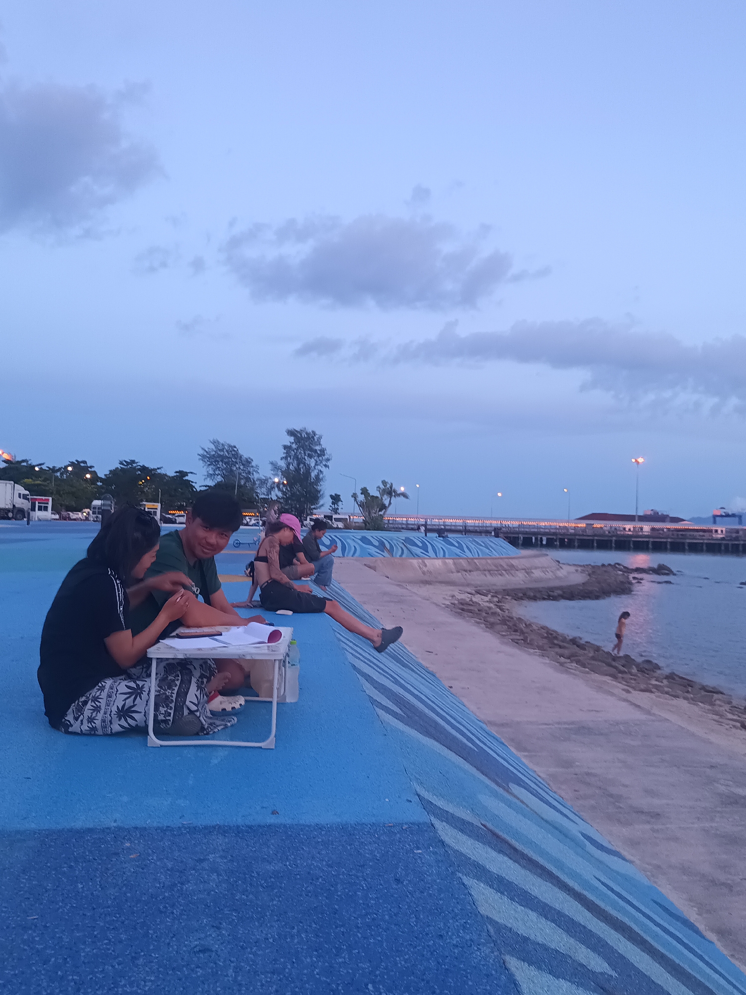 The image depicts an outdoor scene at dusk in Ko Pha Ngan, Thailand. In the foreground, a woman with dark hair is seated on a blue-painted concrete embankment, leaning over a small white table where papers are spread out. She appears to be working on something on the arm of a man sitting next to her, who is looking towards the camera. The man is wearing a dark green t-shirt and has short, dark hair. Behind them, another person in a dark top and a pink hat is seated on the embankment, with a third person sitting further back, appearing to be looking at a phone.
The embankment is painted with a pattern of blue and lighter blue stripes. The sea is to the right of the embankment, with a rocky shoreline. A pier or bridge with illuminated lights stretches across the water in the background, and buildings and trees are visible on the distant shore. A person, possibly a child, is standing near the water's edge on the right side of the image. The sky is a soft twilight blue with scattered clouds. The overall atmosphere suggests a relaxed evening setting by the coast.