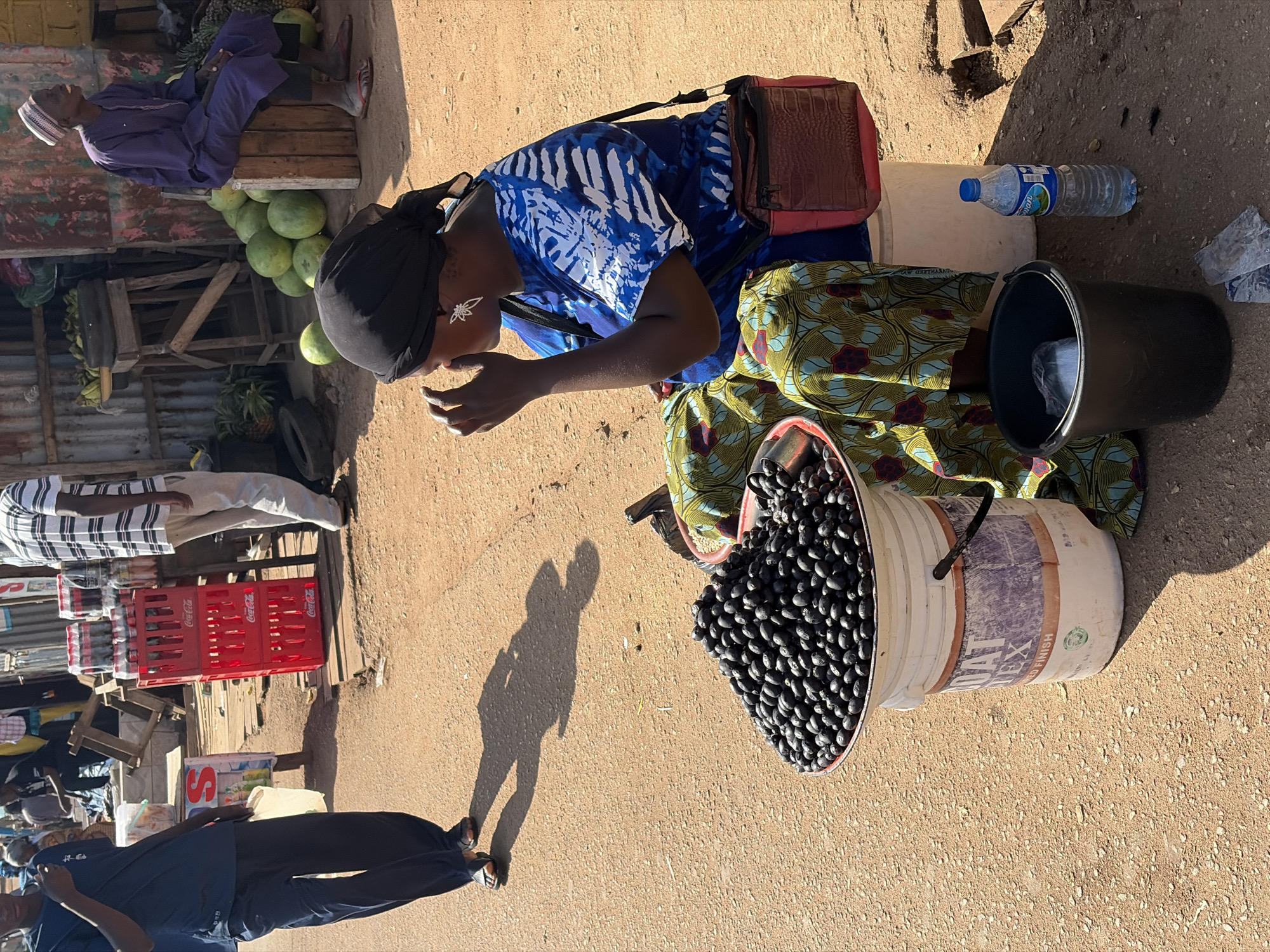 This is a street scene in Jos, Nigeria. In the foreground, a woman with a black headscarf and a blue and white patterned dress sits in front of a large bucket filled with dark, oval-shaped fruits, likely dates or olives. She has a red and brown crossbody bag. To her right, a black bucket and a bottle of water sit on the ground. Behind her, a pile of watermelons and pineapples are visible, along with a person in a purple robe and white cap sitting on a wooden crate. To the left, a person in a navy blue shirt and pants walks away from the camera, casting a shadow on the dusty ground. In the background, crates of Coca-Cola are stacked, and other people are seen in the distance. The environment appears to be an open-air market with stalls and corrugated metal structures.