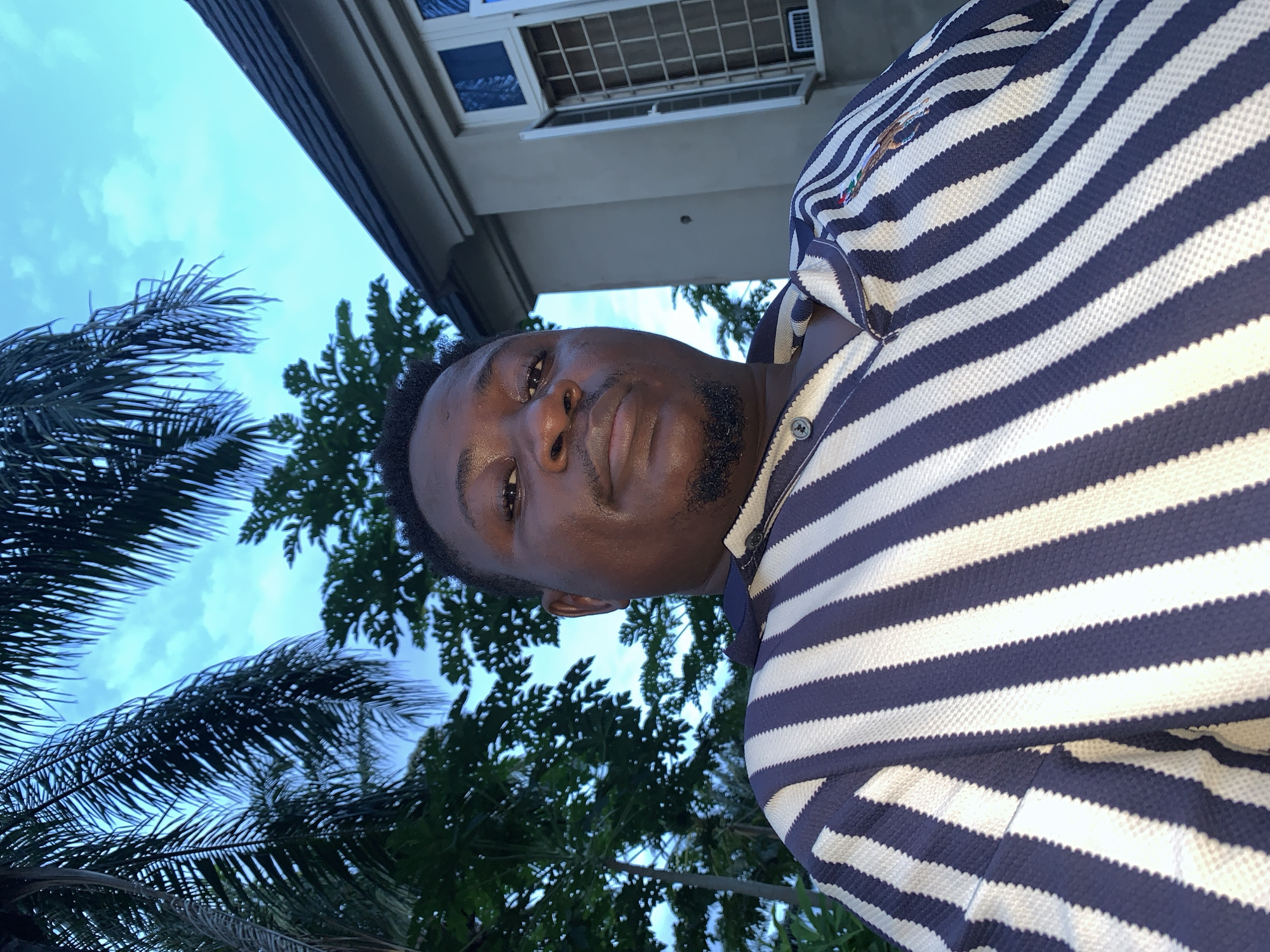 A selfie taken from a low angle shows a dark-skinned man with a goatee and short, dark hair, looking directly at the camera. He is wearing a navy and white striped polo shirt. Behind him, palm fronds and other trees are visible against a bright blue sky with scattered clouds. To the right, a light-colored building with a dark-tiled roof and a window with bars is visible. The scene appears to be outdoors, possibly in a residential area, given the presence of the building and lush vegetation. The lighting suggests it is daytime. The location is identified as Akwanga, Nigeria.