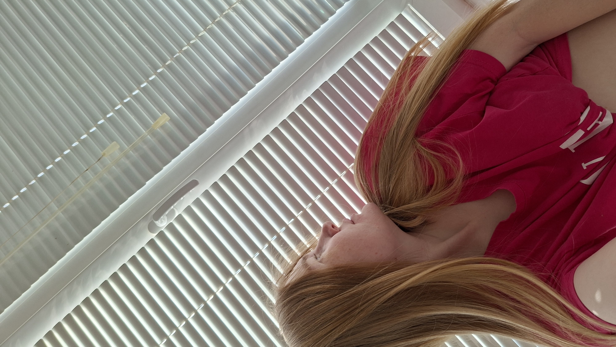 A low-angle, skewed close-up shot depicts a light-skinned person, likely female, with long, blonde hair with pink streaks, lying near a set of white blinds. She wears a magenta-colored top. The blinds, with their horizontal slats, dominate the background, creating strong linear patterns of light and shadow. The person's face is turned slightly to the side, suggesting a relaxed or resting pose. The location is not discernible from the image.