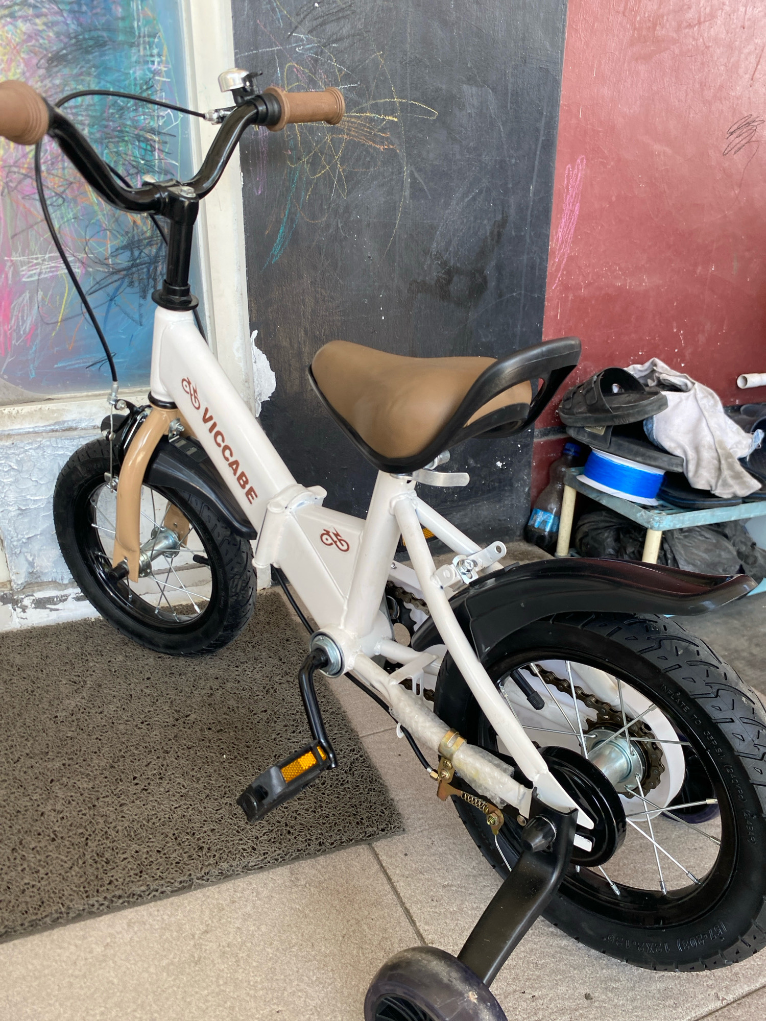 A white children's bicycle with brown handlebars and seat is parked on a gray mat in front of a wall that is half black and half red. The black section of the wall has colorful chalk drawings on it. The bicycle has black tires, a black fender over the rear wheel, and training wheels. The brand name "VICCABE" is visible on the bicycle frame. A small silver bell is attached to the handlebars. In the background, there is a shelf with various items, including a pair of black sandals, a white cloth, a blue spool, and a bottle of dark liquid.