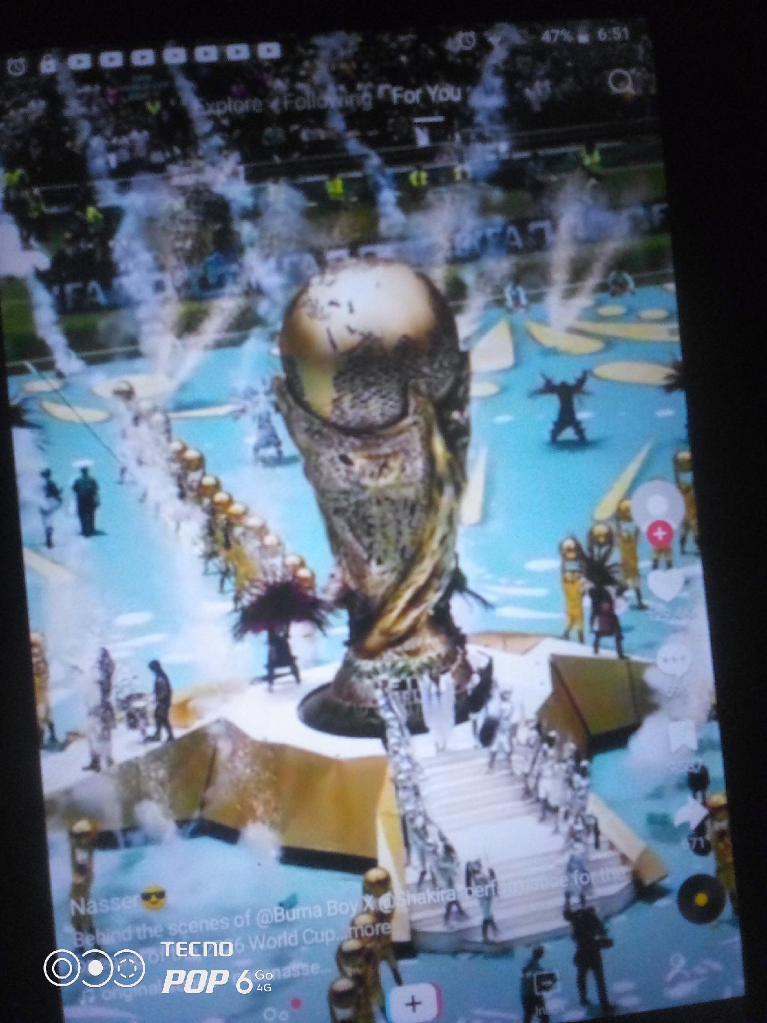 The image displays a screenshot of a TikTok video featuring a virtual rendition of the FIFA World Cup trophy in a stadium setting. The trophy is centrally positioned, surrounded by a blue pool of water and golden structures. There are numerous small, stylized figures scattered throughout the scene, some appearing to be walking or standing. In the foreground, there is text overlay indicating a user named "Nasser" and mentioning "@Burna Boy X @Shakira performance for the World Cup." The bottom of the screen shows the TECNO POP 6 4G logo. The overall scene is digitally rendered, with visible digital artifacts and a low-resolution quality, suggesting it is a mobile phone screen capture of a video. The image does not provide any information about the city of Howayi, Nigeria, nor does it depict any real-world elements from that location.