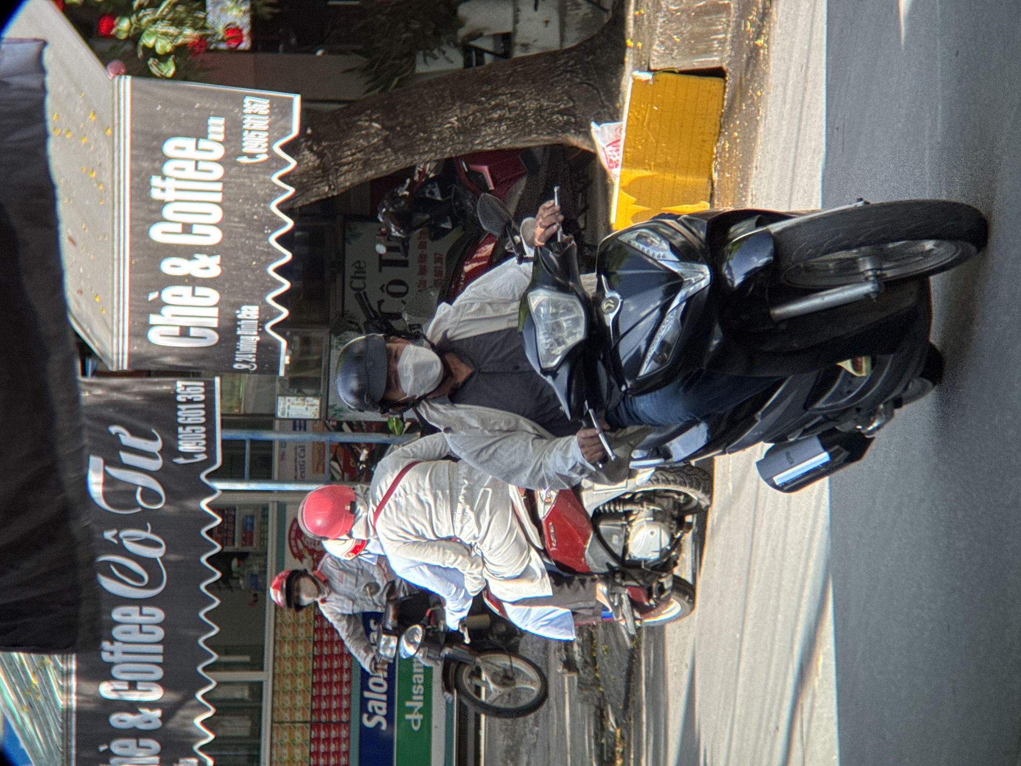The image shows a street scene in Nha Trang, Vietnam. In the foreground, a person is riding a black motorcycle. They are wearing a dark helmet and a white mask. The motorcycle has a silver trim on the front and a black exhaust pipe. Behind this motorcycle, a red motorcycle is visible, with a person wearing a red helmet and a light-colored outfit. Further in the background, another person on a motorcycle is wearing a red helmet and a light grey uniform. Signs for "Chè & Coffee" are visible on the storefronts, with Vietnamese text and phone numbers. The environment appears to be an urban street with buildings and trees lining the road. The lighting suggests it is daytime, with shadows cast on the pavement.