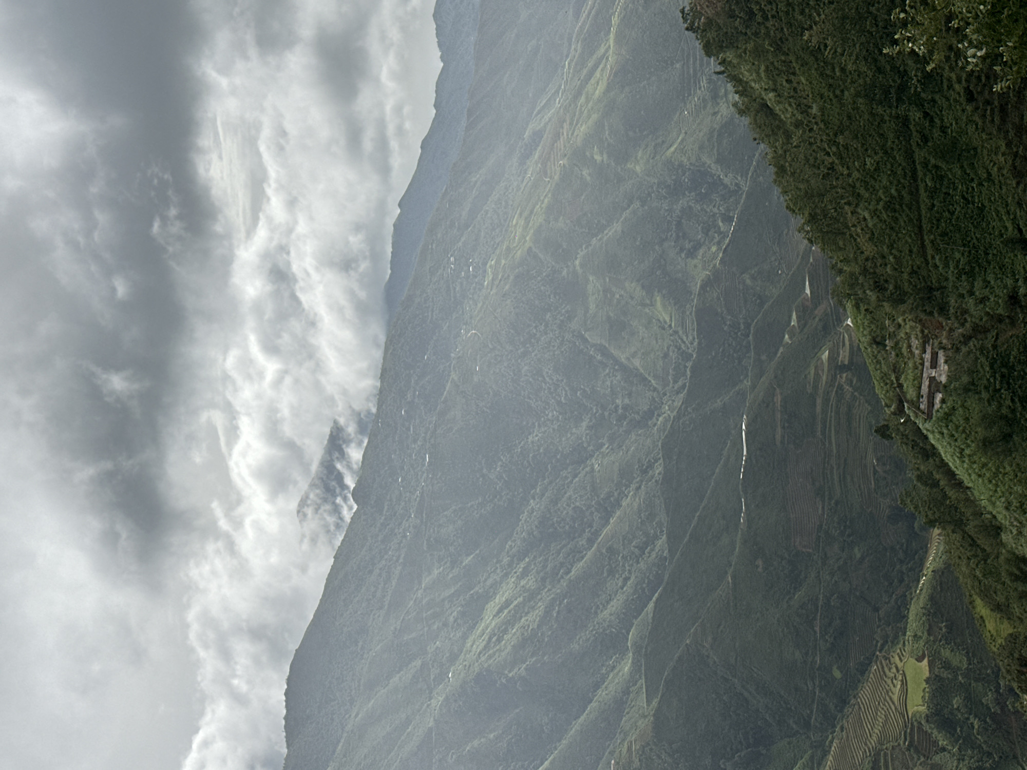 A wide shot captures the undulating, mist-shrouded mountains of Ta Xua, Vietnam. The terrain is characterized by steep slopes covered in a dense, lush green vegetation, suggesting a tropical or subtropical climate. The mountains are terraced, indicating agricultural activity, likely rice paddies or tea plantations, which are visible as geometric patterns carved into the hillsides. Winding roads or paths snake across the landscape, connecting what appear to be small, scattered human settlements nestled within the folds of the mountains. The sky is dramatic and overcast, with heavy, grey clouds casting a diffused light upon the scene, adding a sense of depth and atmosphere. The overall impression is one of rugged natural beauty and rural tranquility.