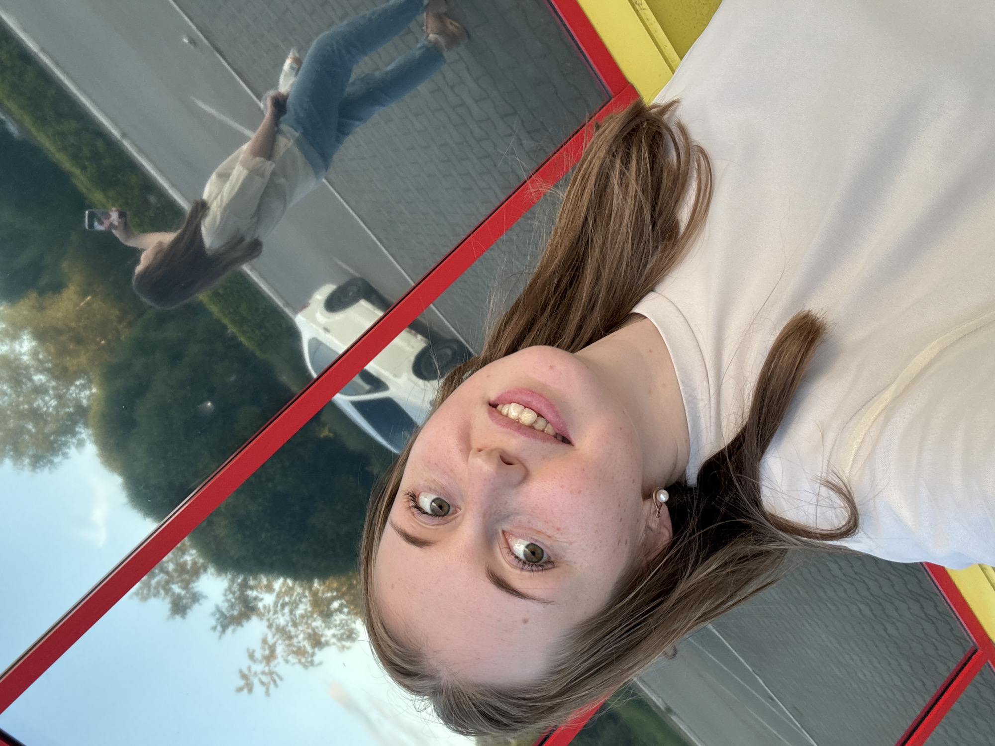 A tilted selfie photograph features a young, light-skinned woman with light brown hair and freckles, appearing from the chest up in the foreground. She wears a light-colored, possibly cream, crew-neck t-shirt and a pearl stud earring on her visible ear. Her eyes, which appear green or hazel, are wide open, and she displays a slight smile showing her teeth, looking directly at the viewer.
The image is captured through a reflective surface, likely a window, indicated by the background elements. In the reflection, the same woman is visible, further back and angled, wearing a light-colored top (matching the foreground subject) and blue jeans. She is actively taking the selfie, holding a smartphone aloft with her right hand, pointed towards herself. A white object is held in her left hand. Behind her, a white car is parked on a paved surface with visible markings. In the upper reflected portion, dark green trees and a light blue sky with faint clouds are visible.
A prominent red structural frame crosses the image diagonally, separating the immediate foreground from the reflected background, with a segment of a yellow frame also visible at the bottom right, suggesting the reflective surface is part of a building or kiosk. The scene is outdoors during daylight in the city of Unknown, Unknown.