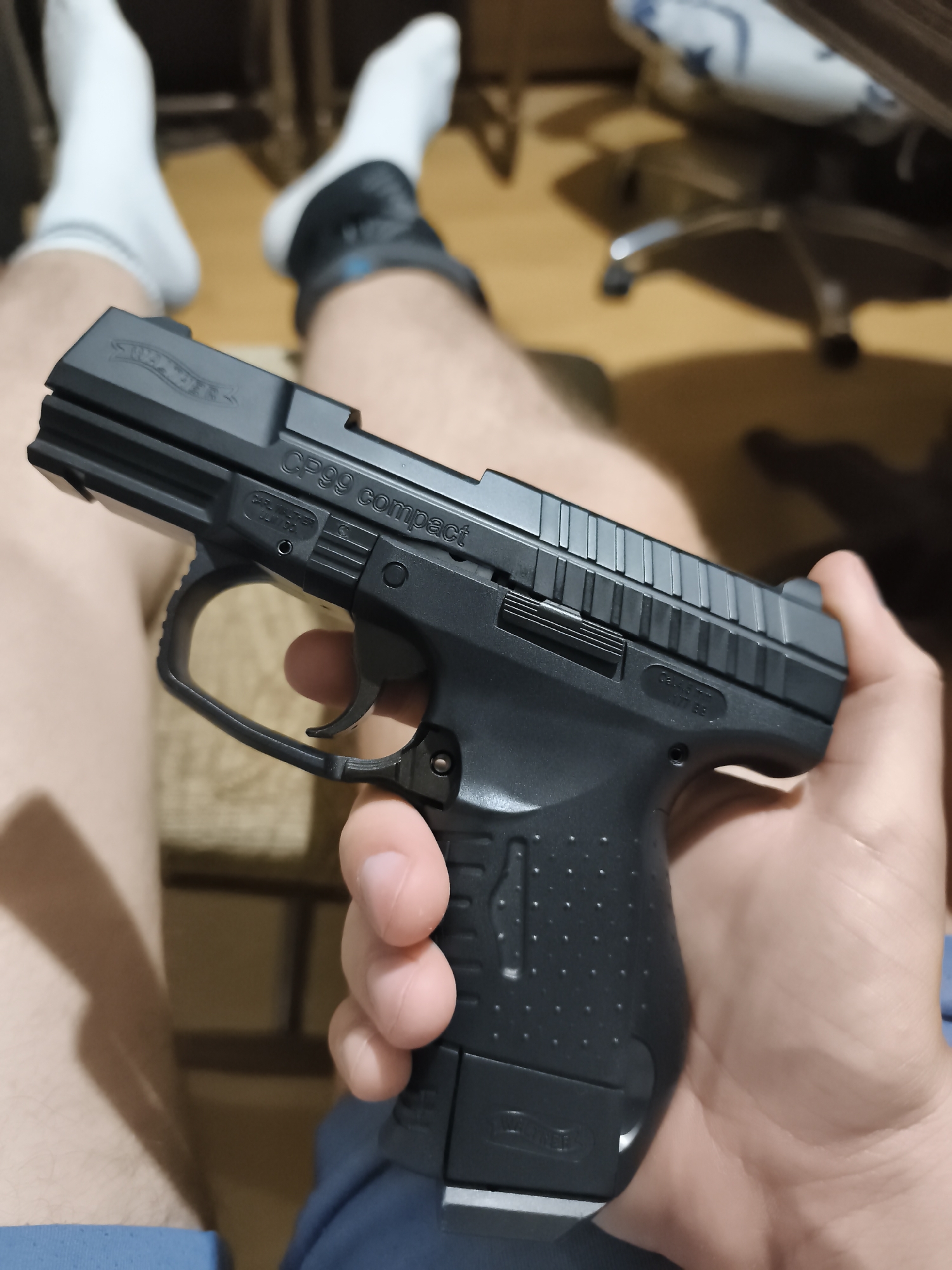An indoor, eye-level shot from Strazhitsa, Bulgaria, shows a person seated with a black pistol in their right hand. The individual's legs, clad in light-colored shorts and white ankle socks (one with a dark band), are visible in the foreground, bent at the knee in a relaxed posture.
The pistol is a black replica with a textured grip and appears to be made of polymer and metal. Clearly visible text on the slide identifies it as a "WALTHER" (with logo) and "CP99 compact". Further text on the frame below the slide reads "CARL WALTHER ULM/DO" and "Cal. 6mm BB", indicating it is likely an airsoft or BB gun. The pistol is held firmly, with the trigger guard around the index finger.
In the blurred background, a light-colored floor is visible, along with parts of a silver-legged office chair on wheels. The lighting suggests an indoor setting, possibly a room with ambient artificial light. No specific time of day or emotions are discernible.