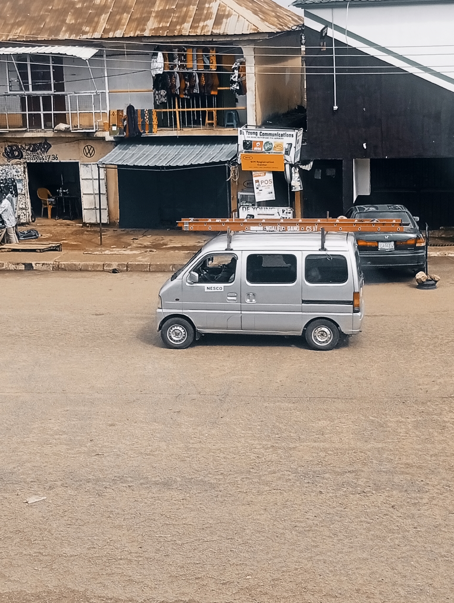 This image depicts a street scene in Bukuru, Nigeria. A silver mini-van with a ladder rack is parked on the dusty road. The van has "NESCO" printed on its side. In the background, there are buildings with various businesses, including what appears to be a communications shop with signs for "DE YOUNG COMMUNICATIONS" and "POS". A dark-colored sedan is also parked in the background. The environment appears to be an urban or semi-urban area, with a slightly weathered appearance to the buildings and road.