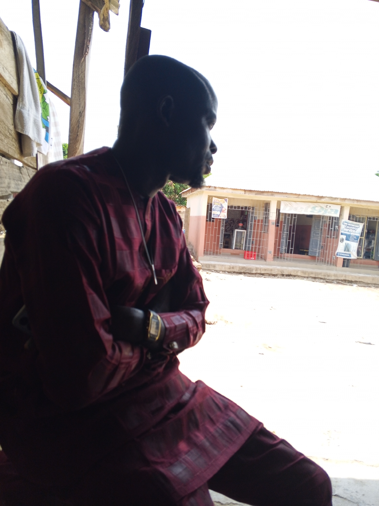 A medium shot depicts a man seated, with his arms crossed, in an open structure, possibly a veranda or covered porch, in Zango-ndaji, Nigeria. The man, dark-skinned, is dressed in a dark red patterned shirt and pants. He wears a watch on his left wrist. A thin silver necklace is visible around his neck. His silhouette is strongly defined due to lighting conditions. The background features a sunlit street with a low, concrete building across from him. The building is rectangular with several storefront windows and advertisements displayed. The sky is bright, suggesting midday. The area is sparsely populated.