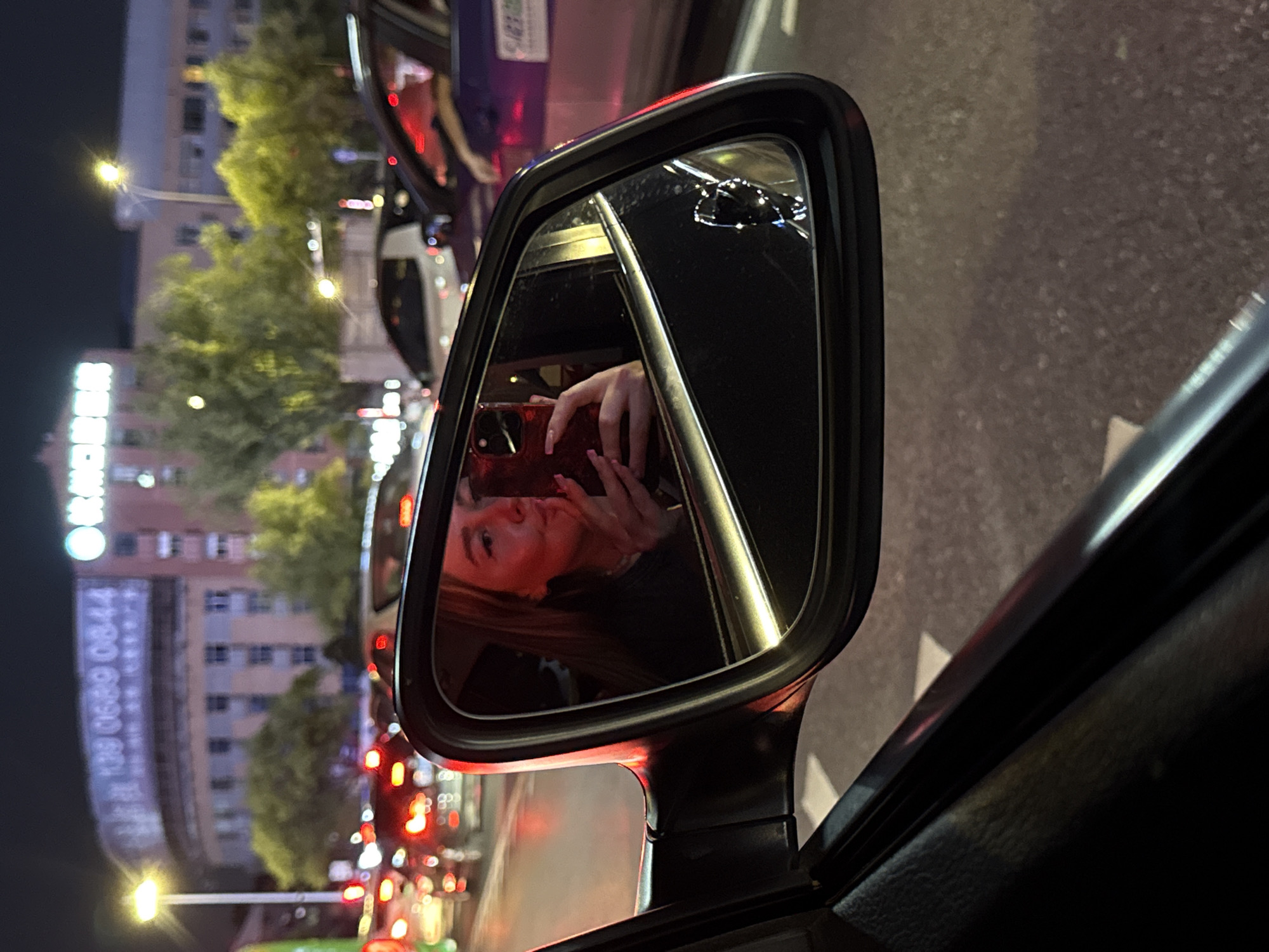 The image depicts a nighttime scene on a city street in Yiwu, China. A car's side mirror reflects a young woman taking a selfie with her smartphone. She is wearing a dark top and has long, blonde hair. Her right hand, adorned with long, pink fingernails, holds the red smartphone. The background in the mirror shows other vehicles and traffic lights, indicating a traffic jam. Outside the mirror, the car's interior is visible, along with the exterior of other cars in traffic. The overall environment is urban, with streetlights and building facades in the distance. A large building with illuminated text that includes Chinese characters and the number "139 0689 0844" is visible in the background, suggesting a commercial or residential area. The atmosphere is typical of a busy city at night.