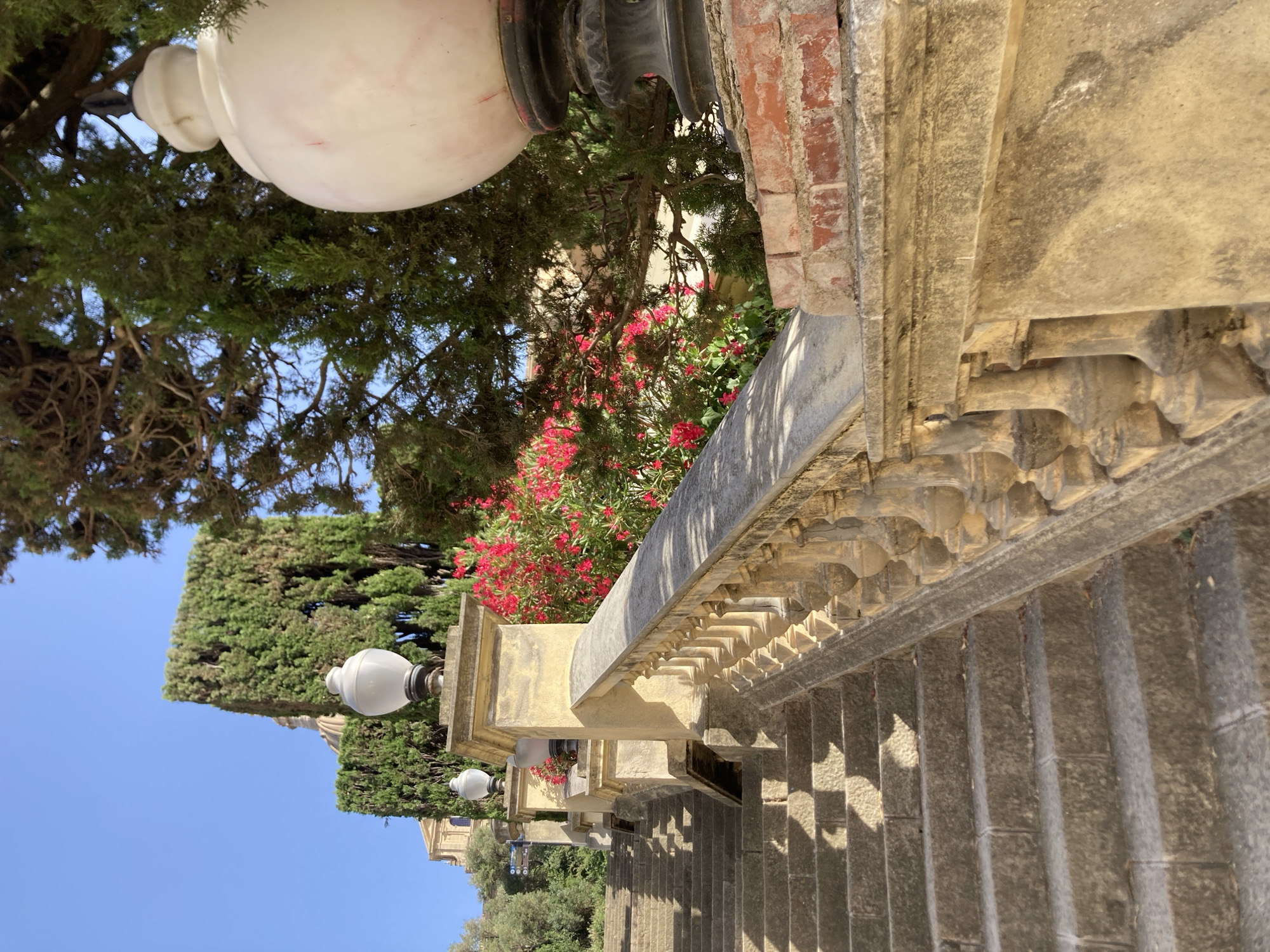 The image depicts an outdoor stone staircase leading upwards. The staircase has a balustrade with ornate stone pillars and a smooth stone railing. Large, spherical lamps are visible at intervals along the balustrade, mounted on stone pedestals. Lush green trees and bright pink flowers form a backdrop to the staircase, with a clear blue sky overhead. In the distance, a building with a domed roof is partially visible. The architecture and landscaping suggest a historical or park-like setting. Based on the architectural style and landscaping, this scene is likely located in the Park Güell in Barcelona, Spain.