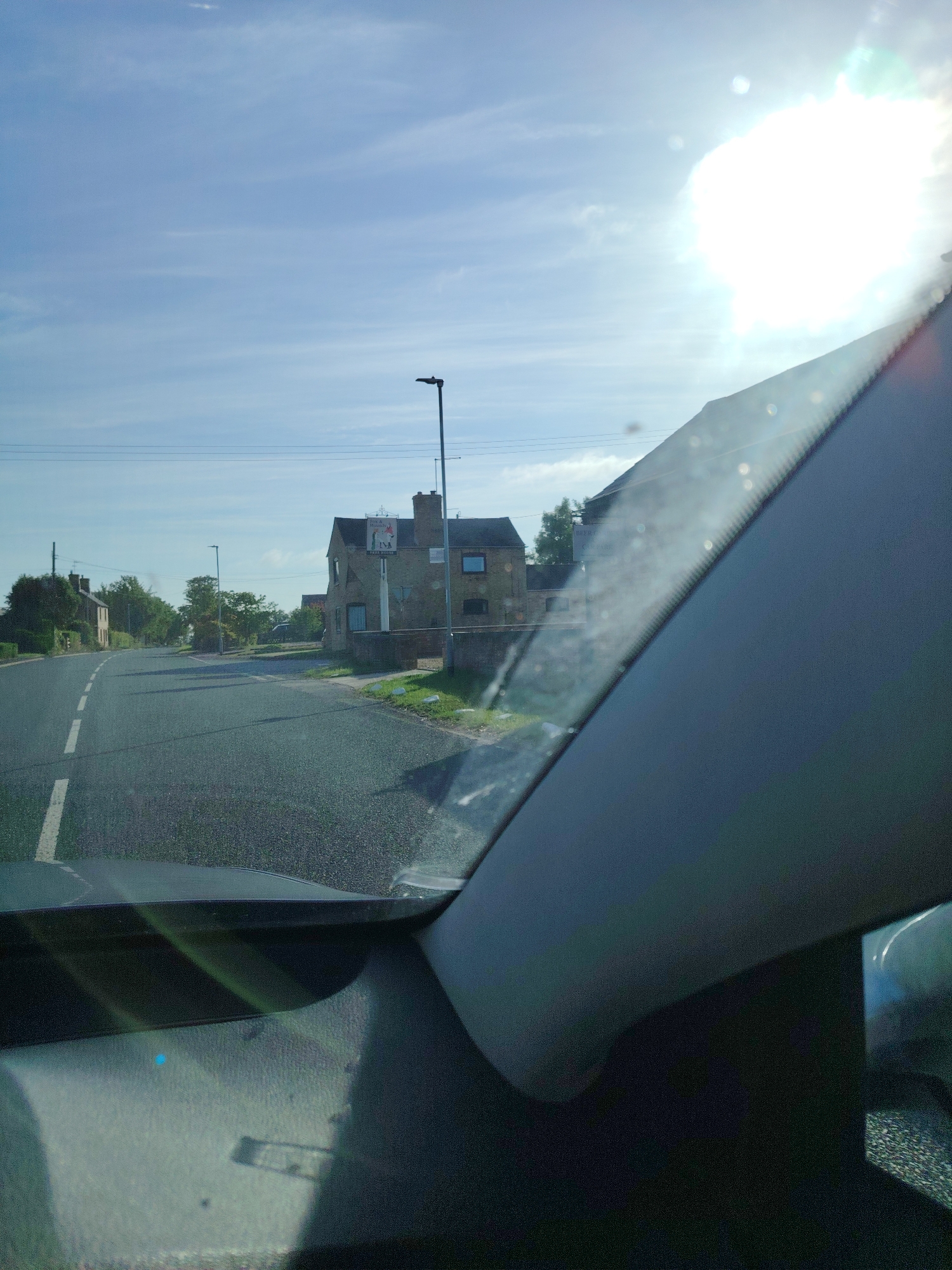 The image presents a daytime scene captured from within a vehicle, looking forward through the windshield onto a rural road in Huntingdonshire, United Kingdom. The bright sun, positioned in the upper right quadrant, causes significant lens flare and casts shadows across the scene.
The immediate foreground displays the dark-toned dashboard and a portion of the passenger-side A-pillar of the vehicle. The windshield shows some reflections and minor dust/smudges.
The road, an asphalt surface with a dashed white centerline, curves gently to the left in the distance. On the right side of the road, a two-story brick building, likely a pub or inn, stands prominent. A sign affixed to the building is partially visible, depicting two figures (possibly a hound and a fox) and text including "Hounds..." and a local Huntingdon (01480) phone number. A tall streetlight is situated near this building.
Further down the road on the left, additional brick residential structures are visible amongst trees and dense foliage. Overhead power lines span across the clear, light blue sky, which features wispy clouds. The environment appears serene, with no active people or other vehicles visible.