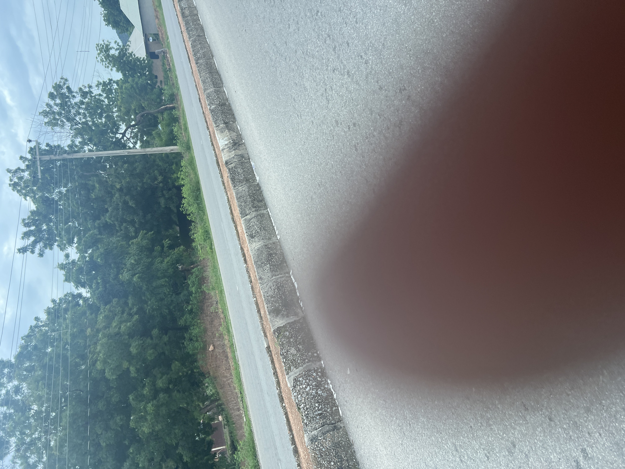The media file shows an outdoor scene with a road and vegetation. A portion of the right side of the image is blurred due to a thumb obscuring the camera lens. The road appears to be paved and has a textured surface. Along the side of the road, there is a curb made of concrete blocks filled with gravel. Beyond the curb, there is a strip of dry earth and then lush green trees. A power pole with wires is visible among the trees on the left side of the road. The sky is partly cloudy, suggesting daytime with moderate weather conditions. The overall setting appears to be a rural or semi-urban area in Kona Garu, Nigeria, given the presence of power lines and the natural vegetation. There is no visible text in the image.