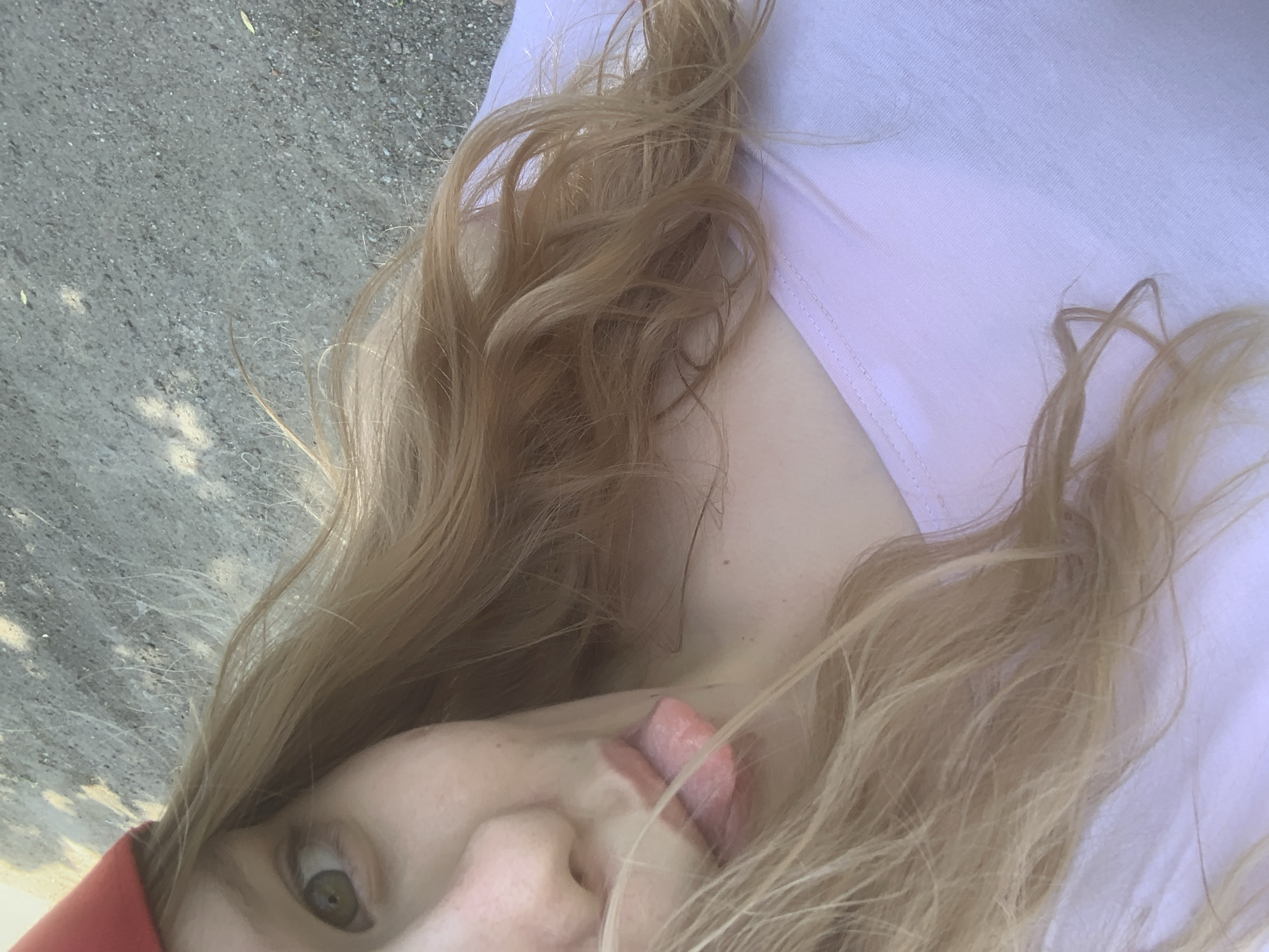 A close-up, high-angle selfie shot captures a person with blonde hair, fair skin, and light-colored eyes, taken in Priozersk, Russia. The image is rotated 90 degrees clockwise, so the subject's head is on the left and their body extends to the right. The person is looking directly at the camera with their left eye wide open, and their tongue is slightly protruding from their mouth, indicating a playful or silly expression.
The subject is wearing a light-colored (possibly white or very pale pink) top, the fabric of which is visible on the right side of the frame. A hint of a red garment or object is visible on the far left, near the subject's head. Their long, wavy blonde hair is disheveled and frames their face, spilling across their neck and the light-colored top.
The background, visible on the upper left side of the image, appears to be a paved surface like asphalt or concrete, suggesting an outdoor setting. The texture of the ground is rough and mottled with varying shades of gray and some lighter specks. The lighting suggests it is daytime, casting soft shadows that are not distinctly visible due to the close-up and high-angle nature of the shot. The overall impression is an informal and candid moment. No visible text is present in the image.