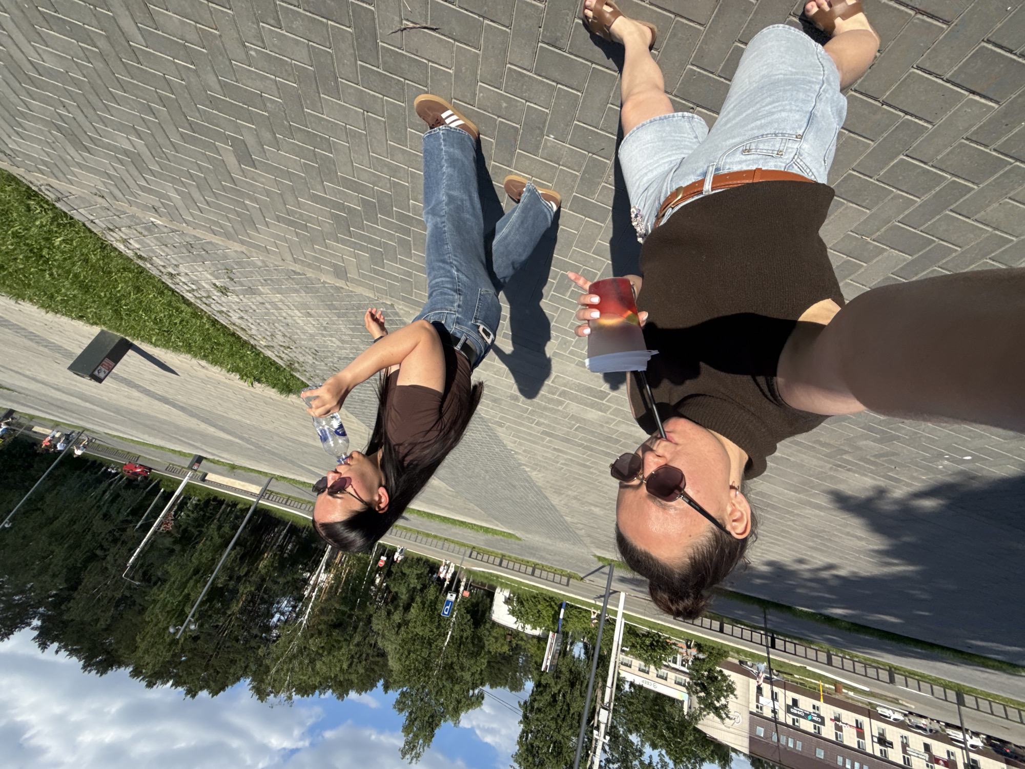Two women are depicted on a paved pedestrian walkway on a sunny day in the city of Chaikovsky, Russia. The woman on the left, appearing to be taking a selfie, is positioned closer to the viewer. She has dark hair styled in a bun, wears dark sunglasses, a dark brown sleeveless knit top, light blue denim shorts, a brown leather belt, and brown leather sandals. Her left arm is extended towards the camera, and her right hand holds a transparent plastic cup containing a reddish beverage with ice and fruit slices, from which she sips through a dark straw. Her shadow is cast diagonally forward onto the paving.
The second woman, to the right and slightly behind the first, is in mid-stride, walking in the same general direction. She has long dark hair, wears dark sunglasses, a dark brown short-sleeved t-shirt, dark blue jeans with a dark belt featuring a silver buckle, and brown sneakers with white stripes (resembling Adidas Gazelles). She is drinking from a clear plastic bottle of "ХВАЛОВСКАЯ" (Khvalovskaya) branded water.
The ground is paved with gray rectangular bricks arranged in a herringbone pattern. A narrow strip of green grass separates the paved area from an asphalt road visible in the middle ground, where white and red cars are seen. In the background, numerous tall trees, including birches and conifers, form a dense wooded area or park. Several streetlights and poles are distributed along the road. In the far left background, multi-story buildings are visible, one being brown/beige with multiple windows. A prominent "СБЕР" (SBER) sign, indicative of a Sberbank branch, is visible on a building further back, confirming a Russian location. The sky overhead is blue with scattered white cumulus clouds. Bright sunlight casts distinct shadows, indicating daytime.