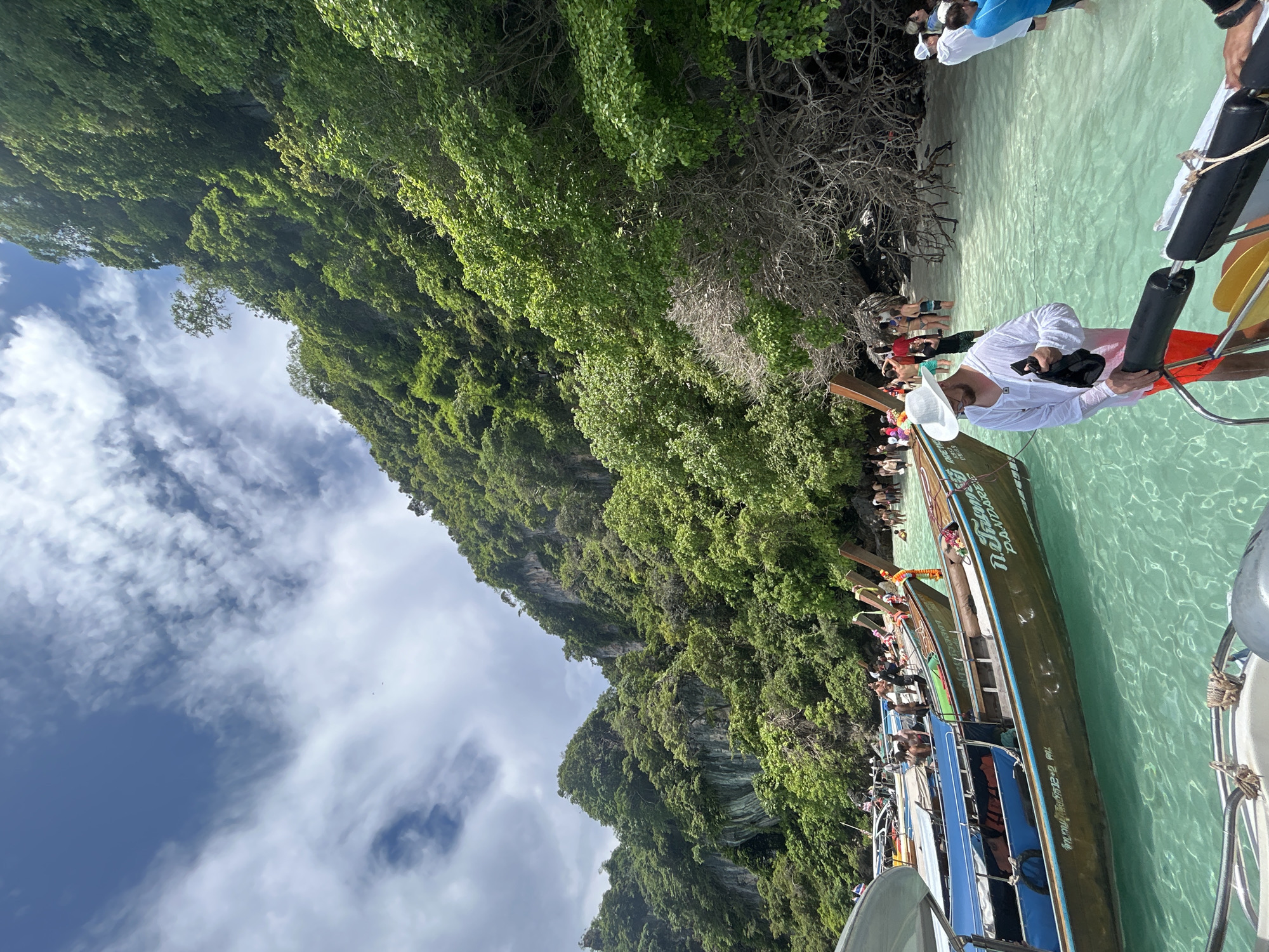 The image depicts a sunny day at a beach in Ko, Thailand, likely on Phi Phi Island, known for its limestone cliffs and clear turquoise waters. In the foreground, a man wearing a white hat and white long-sleeved shirt over orange swim shorts stands on the edge of a boat, holding a black bag. The boat is a traditional longtail boat, with Thai writing visible on its side. Several other longtail boats are moored in the shallow, crystal-clear water.
In the background, a lush green, heavily forested cliff face rises steeply from the water. The beach is populated by numerous people, some wading in the water, others standing near the boats or walking along the shore. The sky is partly cloudy with bright white clouds against a blue backdrop. The overall scene conveys a vibrant tropical tourist destination.
