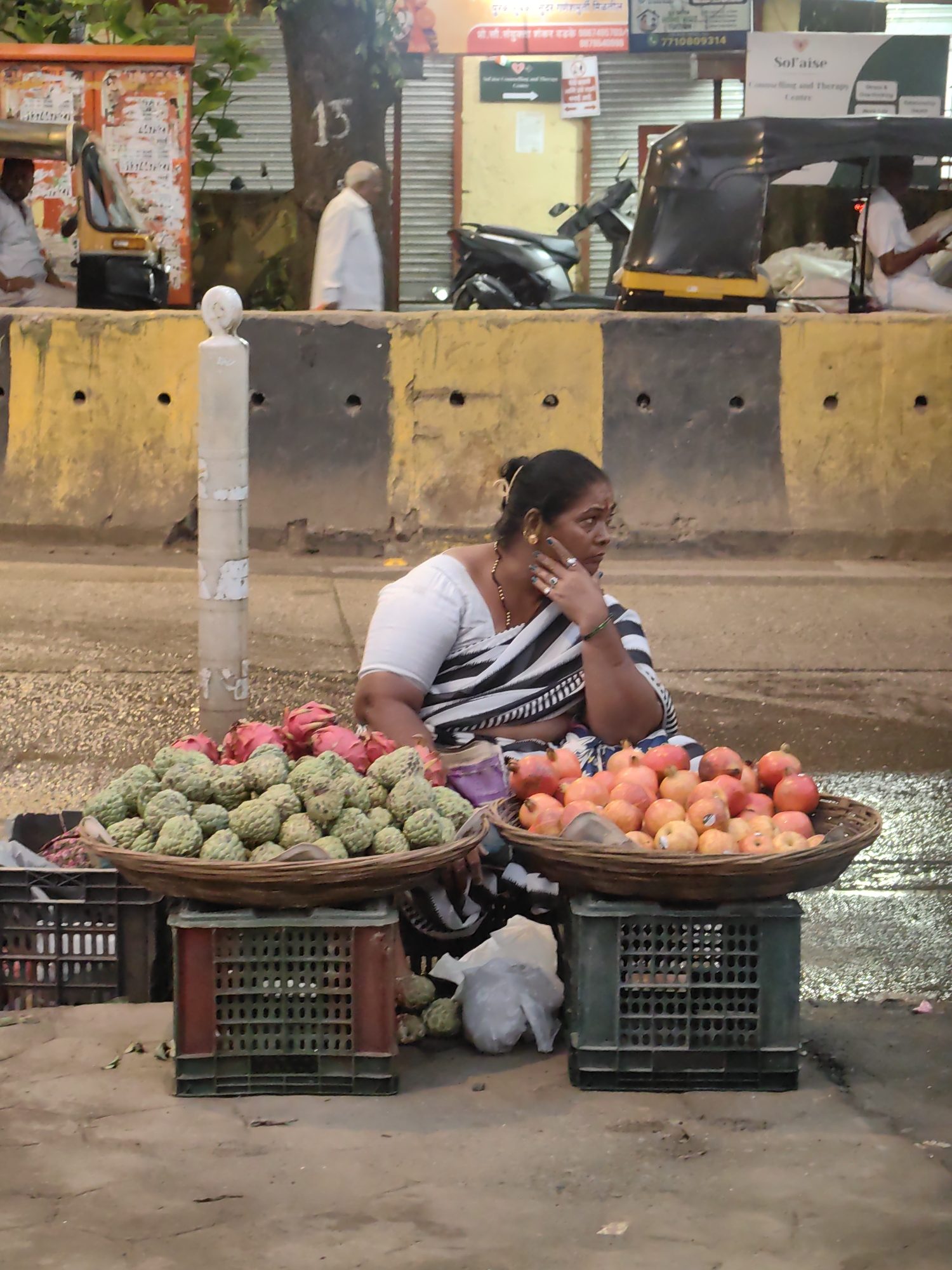 The image depicts a street scene in Mira Bhayandar, India, at dusk. A woman in a black and white striped saree sits by a roadside fruit stall, with her hand resting on her chin. She appears to be selling fruit from two woven baskets placed on top of stacked plastic crates. The front basket contains a large quantity of custard apples, while the basket behind it holds red-skinned fruits, likely dragon fruit. Another basket in front of the woman contains what appear to be pomegranates and other smaller round fruits. The background shows a yellow and black concrete barrier, a tree with the number "13" painted on its trunk, and an auto-rickshaw. A scooter and a building with signage are also visible in the distance. The ground is wet, suggesting recent rain.