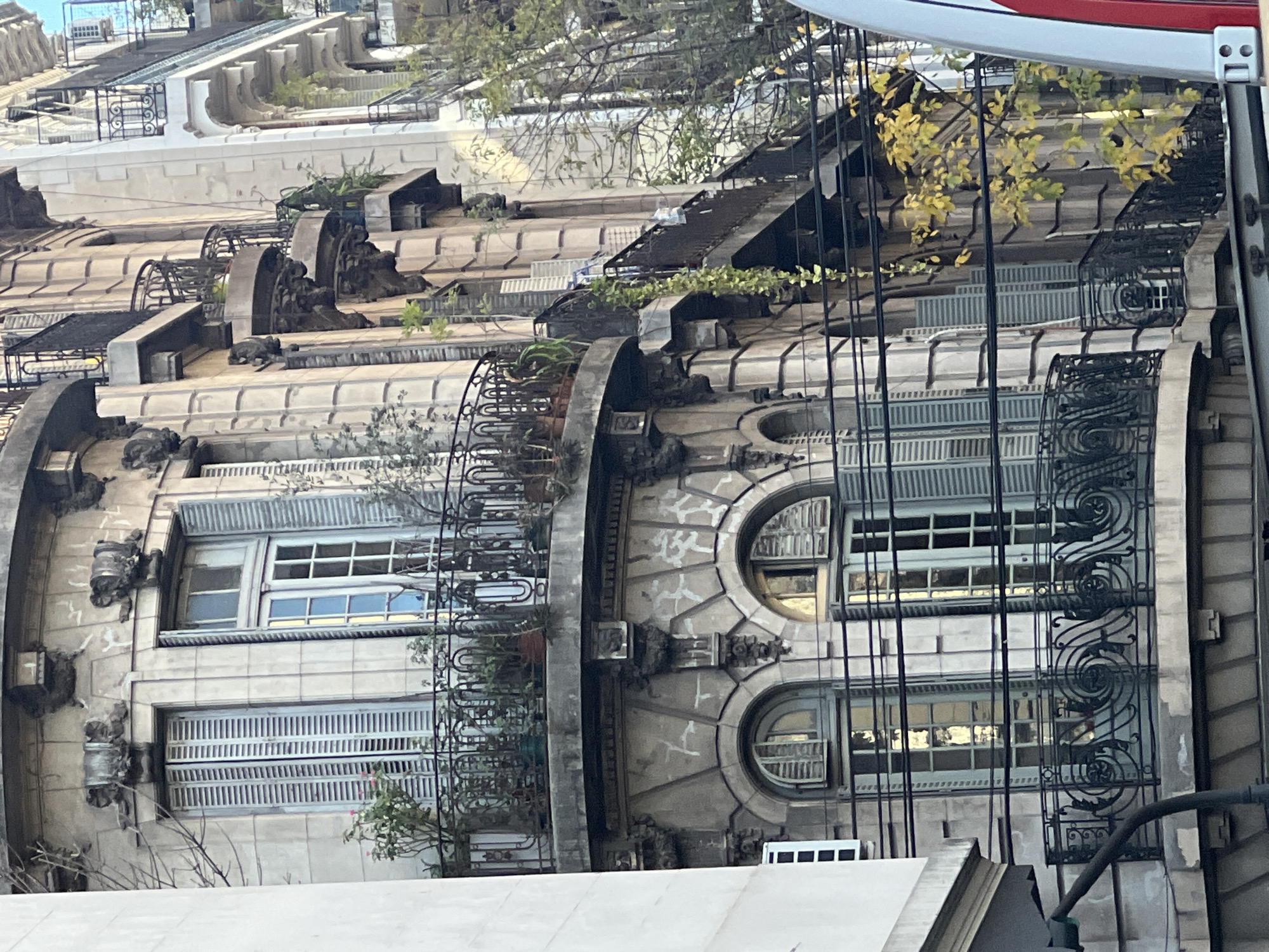 The image shows a section of the Palacio Barolo building in Buenos Aires, Argentina. The architecture features elaborate, multi-tiered facades with ornate ironwork balconies, arched windows, and decorative stonework. Plants and greenery are visible on the balconies. The building's cream-colored exterior is weathered. A red and white object is partly visible at the top-right edge of the frame. Utility lines are in the foreground.