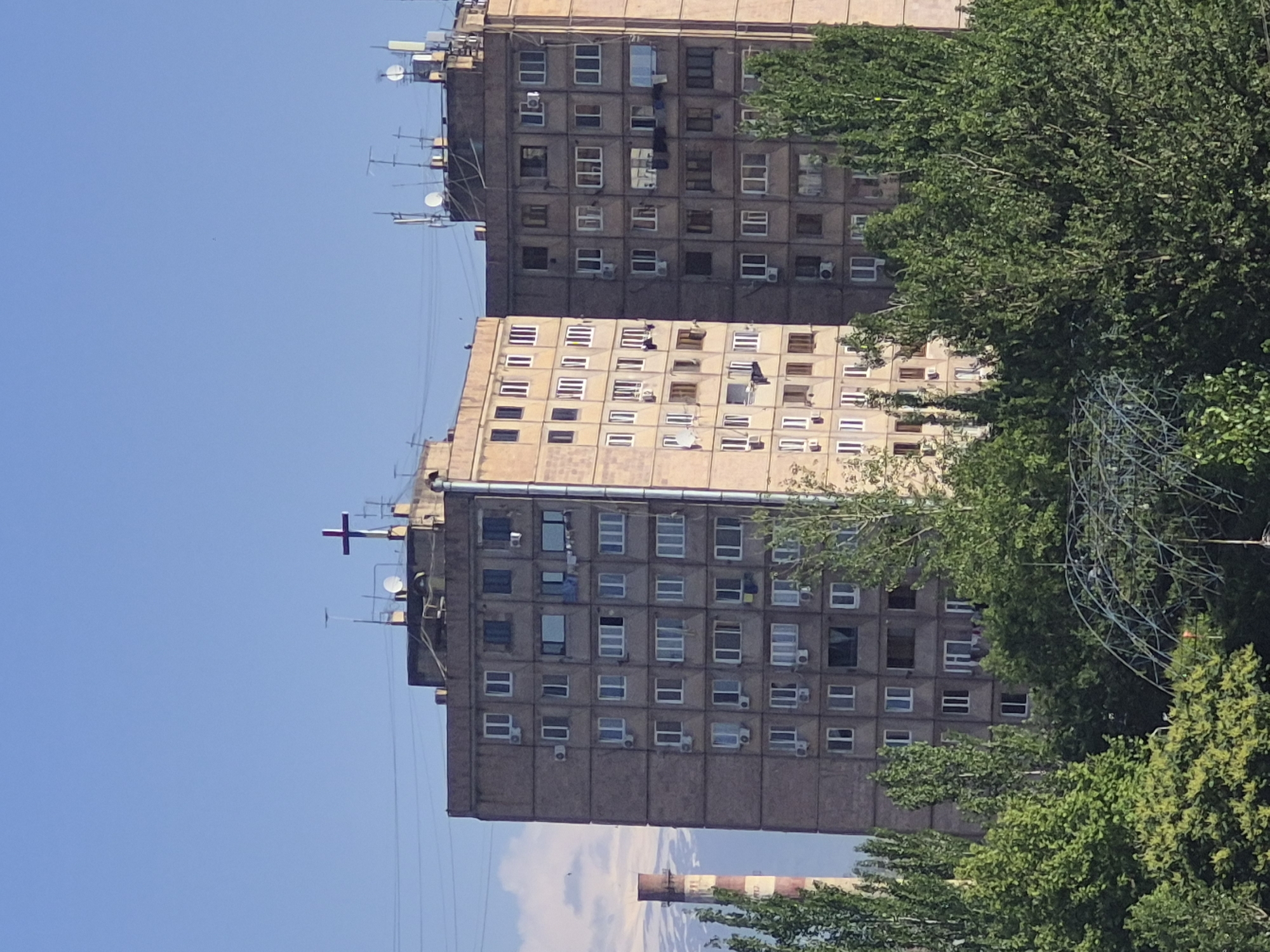 The image, rotated 90 degrees clockwise, displays a daytime scene dominated by two large, multi-story residential buildings, likely apartment blocks, set against a clear blue sky. Both buildings appear to be constructed from concrete or a similar material, with numerous square and rectangular windows, many featuring white frames. Air conditioning units are visible mounted below several windows on both structures, indicating individual apartment installations.
The building in the foreground, slightly to the left, is topped with a distinct red cross, possibly a religious symbol or part of a communication tower. This building also features several antennas and satellite dishes on its roof. The building behind it, to the right, is partially obscured by trees but shows a similar architectural style and rooftop equipment.
Lush green trees and dense foliage frame the buildings on the right side and extend into the foreground, with some branches obscuring parts of the facades. A section of what appears to be a wire mesh structure or part of a park/playground, also covered in greenery, is visible among the trees on the right.
In the far background, below the main buildings, a tall, striped industrial chimney or smokestack, red and white in color, is partially visible against the sky, suggesting an industrial presence nearby. Power lines or cables are faintly discernible stretching across the blue sky between the buildings. The bright lighting and clear sky indicate a sunny day. No people or visible text are present in the image. The scene captures an urban landscape consistent with the provided location context of Yerevan, Armenia.