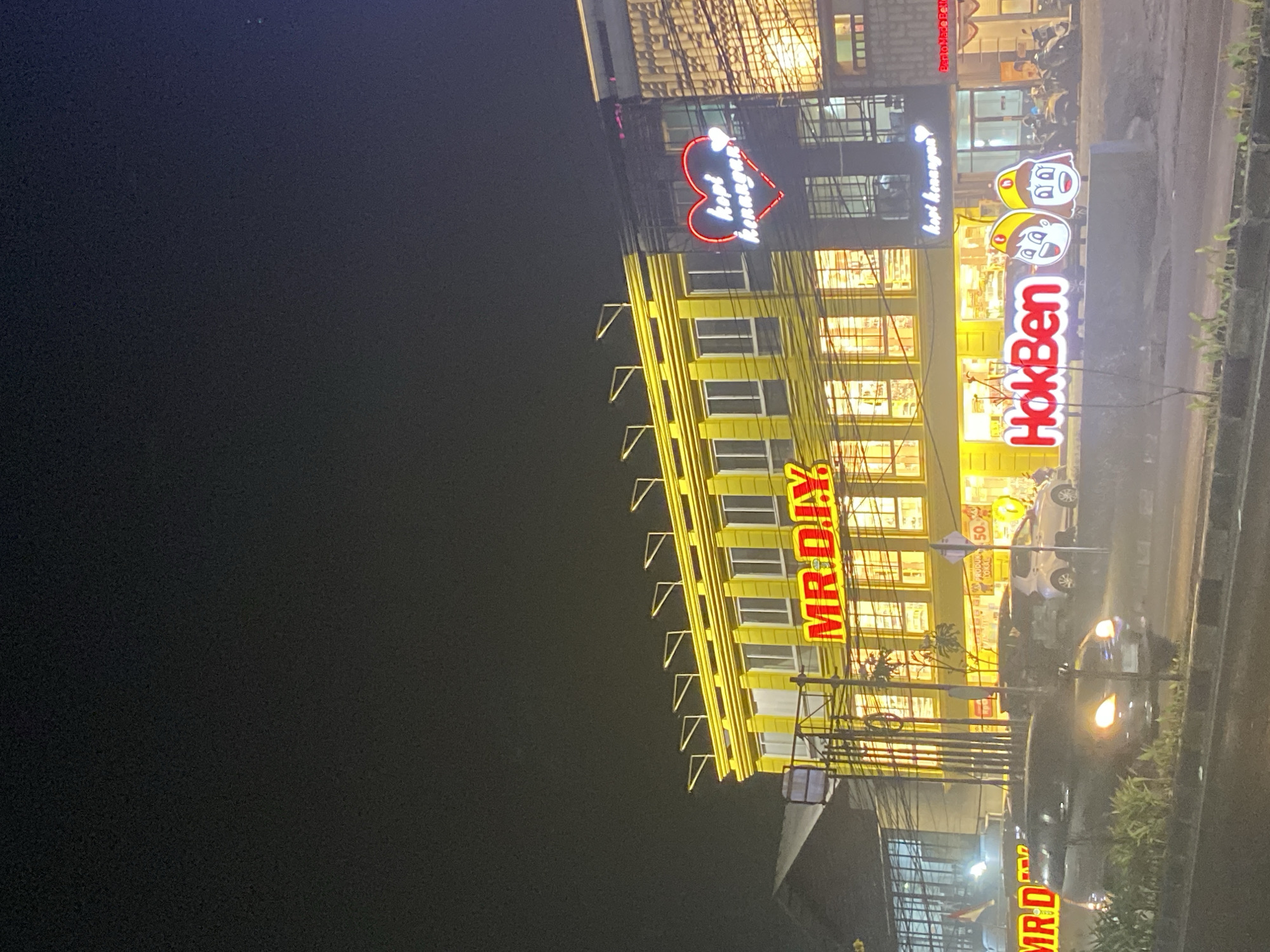 The image shows a night scene in Tangerang, Indonesia, focusing on a brightly lit row of businesses. The most prominent building is a yellow structure with "MR. D.I.Y." displayed in large red neon letters across its facade. Above the windows, a series of yellow triangular lights are mounted. To the right of the MR. D.I.Y. store, another building has a large red neon heart with "kopi kenangan" written in white neon inside, indicating a coffee shop. Further to the right, more storefronts are visible, illuminated with warm interior lights. In front of the businesses, a large neon sign for "HokBen" is displayed, featuring cartoon mascots of a boy and a girl. Several cars and motorcycles are parked or driving on the road in front of the buildings. The overall environment appears to be a commercial street at night, with clear signage and the glow of artificial lights against the dark sky.