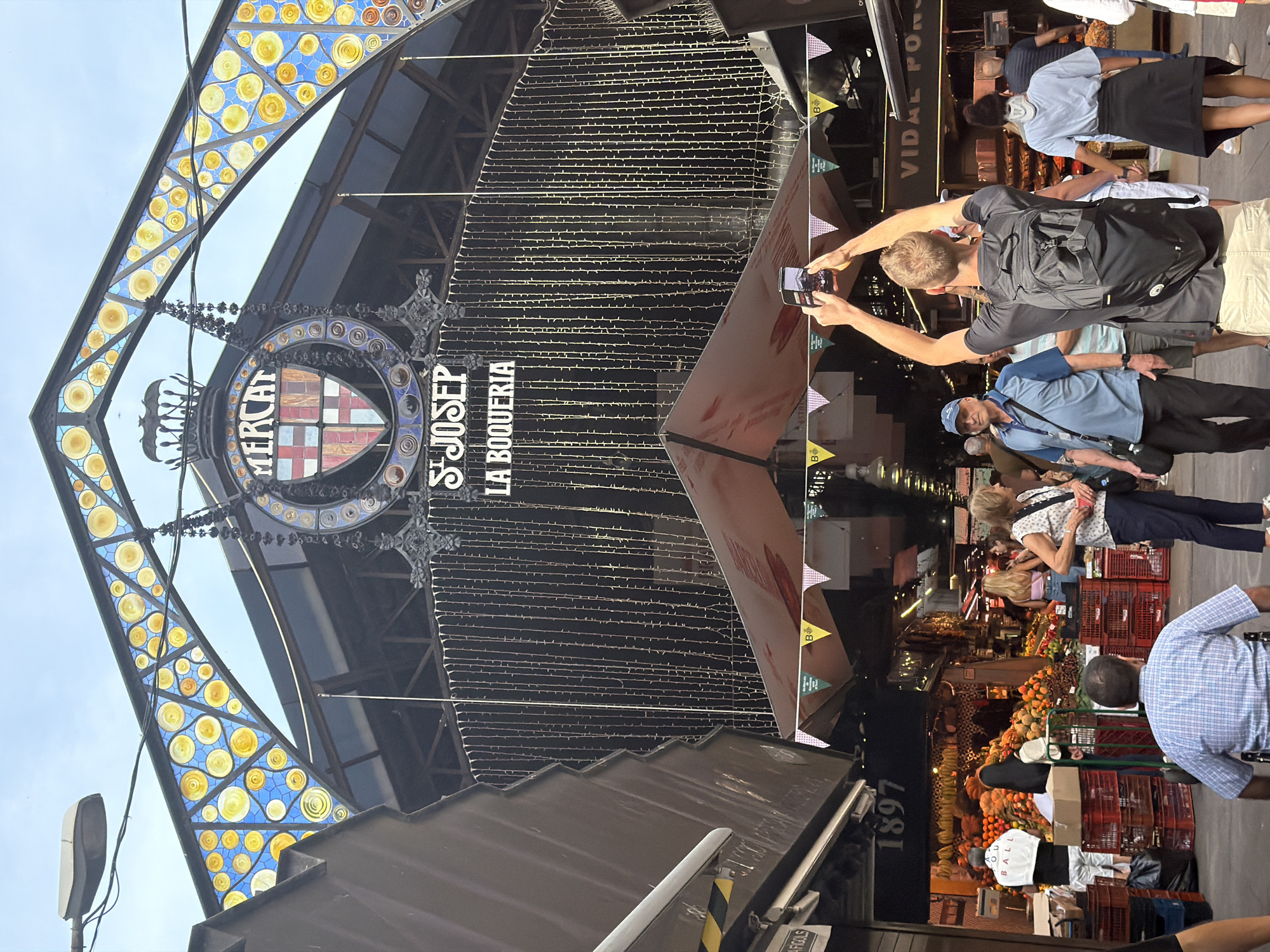 The image shows the entrance to a market. Above the entrance, a large sign reads "MERCAT ST JOSEP LA BOQUERIA". The entrance is adorned with decorative arches and stained glass elements. A person in the foreground is holding up a phone to take a picture of the market entrance. Several other people are visible in the market, browsing at stalls laden with produce. The market appears to be outdoors, with a covered roof structure over the stalls. The location is identified as the city of l'Hospitalet de Llobregat, Spain.
