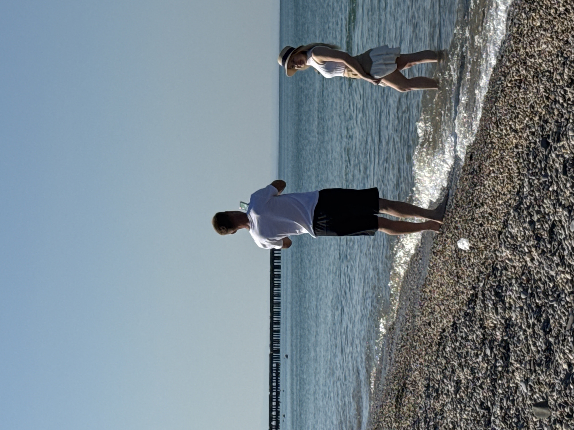 A man stands on a pebble beach in Novofedorovka, Ukraine, holding a phone up to take a picture of a woman. The woman is standing in the shallow water, facing the man. She is wearing a white top, a straw hat, and a white skirt. A pier is visible in the background, extending into the calm blue sea. The sky is clear and blue.