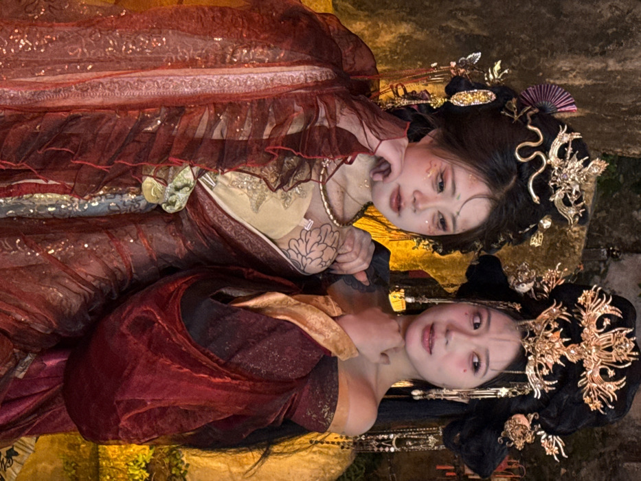 Two East Asian females are depicted in a vertical, waist-up, illuminated shot, situated against a dark, textured background with prominent warm, golden lighting. The scene is located in Yuzhong District, China.
The female on the left has dark hair styled high with an elaborate gold-colored headpiece featuring a serpent-like design and a purple fan motif. Her face displays reddish eye makeup, gold glitter on her cheeks and forehead, and small decorative dots near her outer eyes and temples. She wears a multi-layered, sheer reddish-brown gown with sparkling accents. A black ink floral tattoo is visible on her upper left arm. Her right hand is positioned near her chin, and her gaze is directed downward and to her left.
The female on the right has dark hair styled with an ornate gold-colored headpiece featuring a central phoenix or bird motif and dangling pearl-like ornaments. Her makeup includes reddish eye accents and small red heart-shaped marks below her eyes. She is dressed in a rich, off-the-shoulder maroon and gold-trimmed gown with voluminous drapery. Her left arm is extended, appearing to rest on the left subject's shoulder, while her left hand is near her own chin/neck. She looks to her right with a subtle smile.
The background consists of dark, uneven surfaces resembling rock or stone, with intense artificial golden light illuminating sections behind and to the lower right of the subjects, creating a dramatic contrast. The interaction between the two subjects is posed.