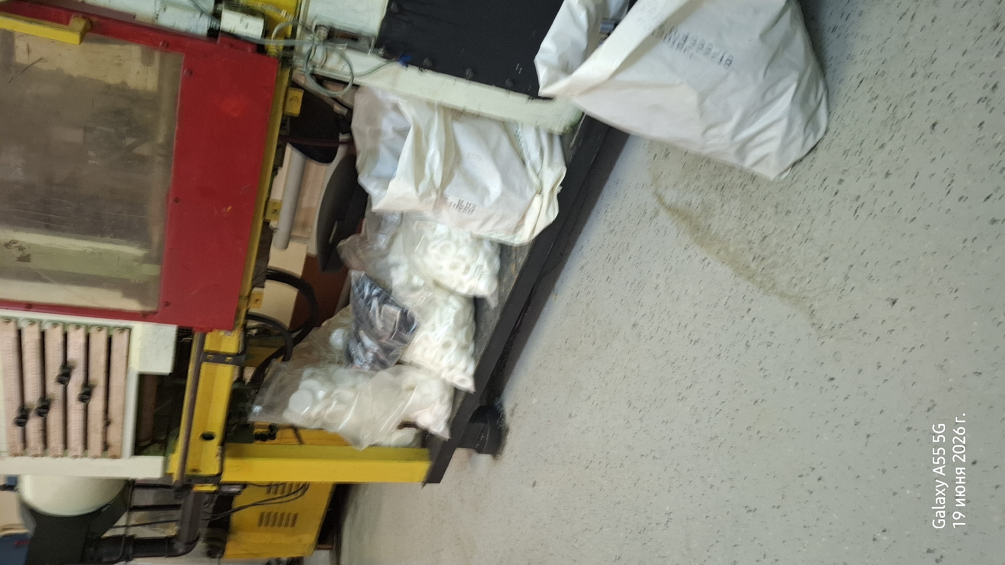 The image depicts an industrial setting with a piece of machinery in the background. In the foreground, several white plastic bags filled with small white ring-shaped objects are arranged on a black platform. The floor is a speckled light gray. A portion of a white bag is visible in the lower right corner, and text is present in the lower left corner. The environment appears to be a warehouse or factory.