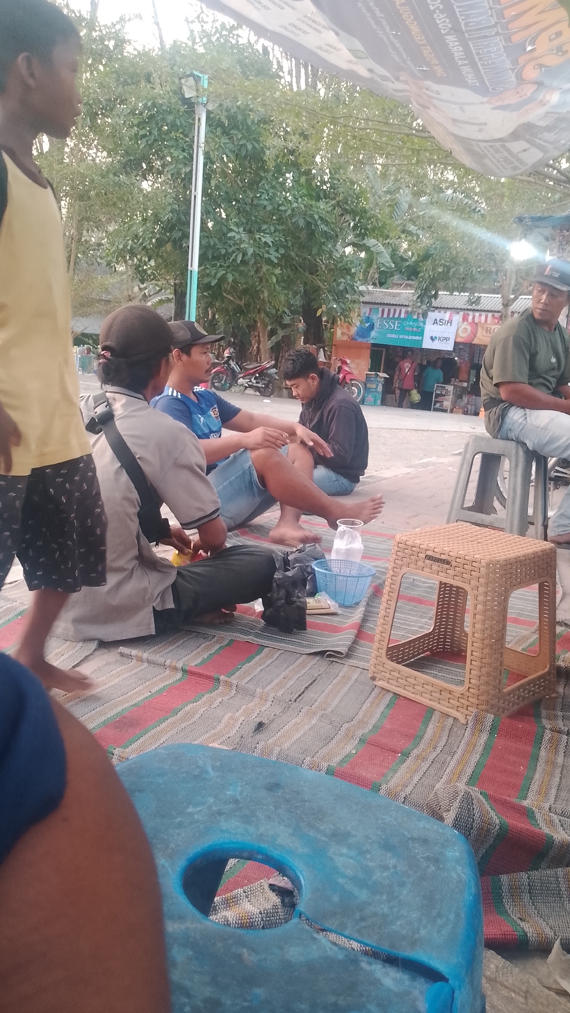 In the city of Karanglangu, Indonesia, a group of men are gathered on a striped mat on the ground, possibly enjoying a casual meeting or meal. The environment suggests a semi-outdoor setting, with trees and buildings visible in the background.
In the foreground, a young boy in a yellow shirt and patterned shorts is partially visible, walking away from the camera. To his left, a man wearing a grey shirt and dark pants, with a dark cap, is seated. He is engaged in an action, possibly preparing something or handling items on the mat. Next to him, a man in a blue and white striped t-shirt and denim shorts sits with his legs crossed. He is looking towards the right. To their right, another man in a dark hoodie is seated, looking down.
Further into the background, several elements are visible. A man in a green shirt and jeans is seated on a plastic stool to the right. Behind the seated men, there are shops or stalls with signage, including one that reads "ESSE DOUBLE UP ENJOYMENT". Motorcycles are parked in the background, indicating a public or accessible area. A light-colored, woven plastic stool is placed on the mat. A blue plastic object with a hole in the center is in the lower left corner of the frame, partially obscuring the ground and mat. A light blue plastic basket is also visible on the mat, containing white packets. Several black plastic bags are scattered on the mat. The overall scene depicts a relaxed social gathering in a vibrant, possibly commercial, area of Karanglangu.