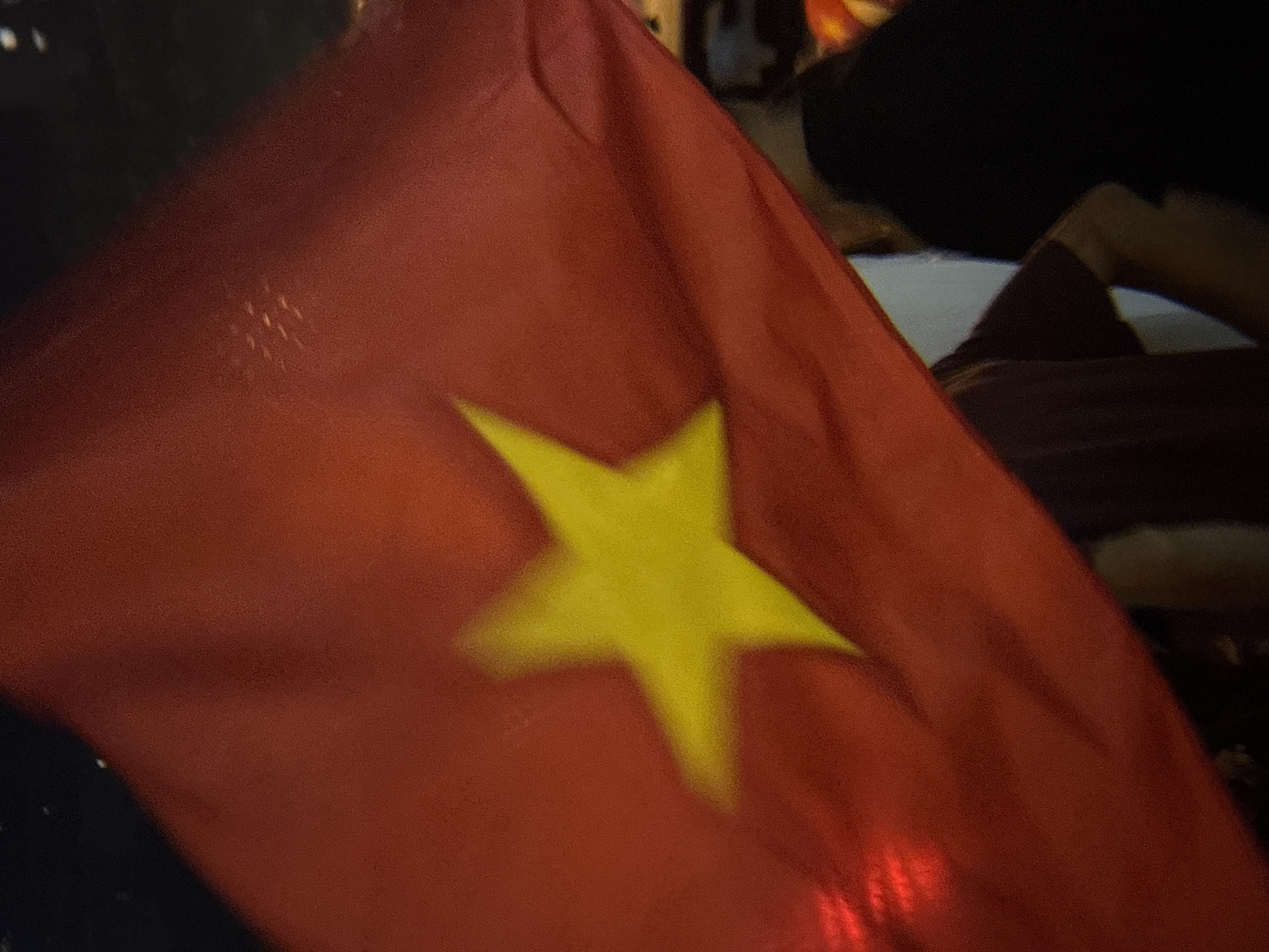 The image shows a close-up of a red flag with a yellow star. The flag appears to be waving, as evidenced by the ripples and folds in the fabric. In the background, several blurred figures are visible, suggesting that the flag is being held by someone in a crowd. The lighting is dim, with some red and yellow reflections on the flag, and the background is mostly dark. There is no clear indication of the specific location within Nha Trang, Vietnam, or any specific action or interaction taking place. The image is a still photograph of a flag, with people and the environment out of focus.