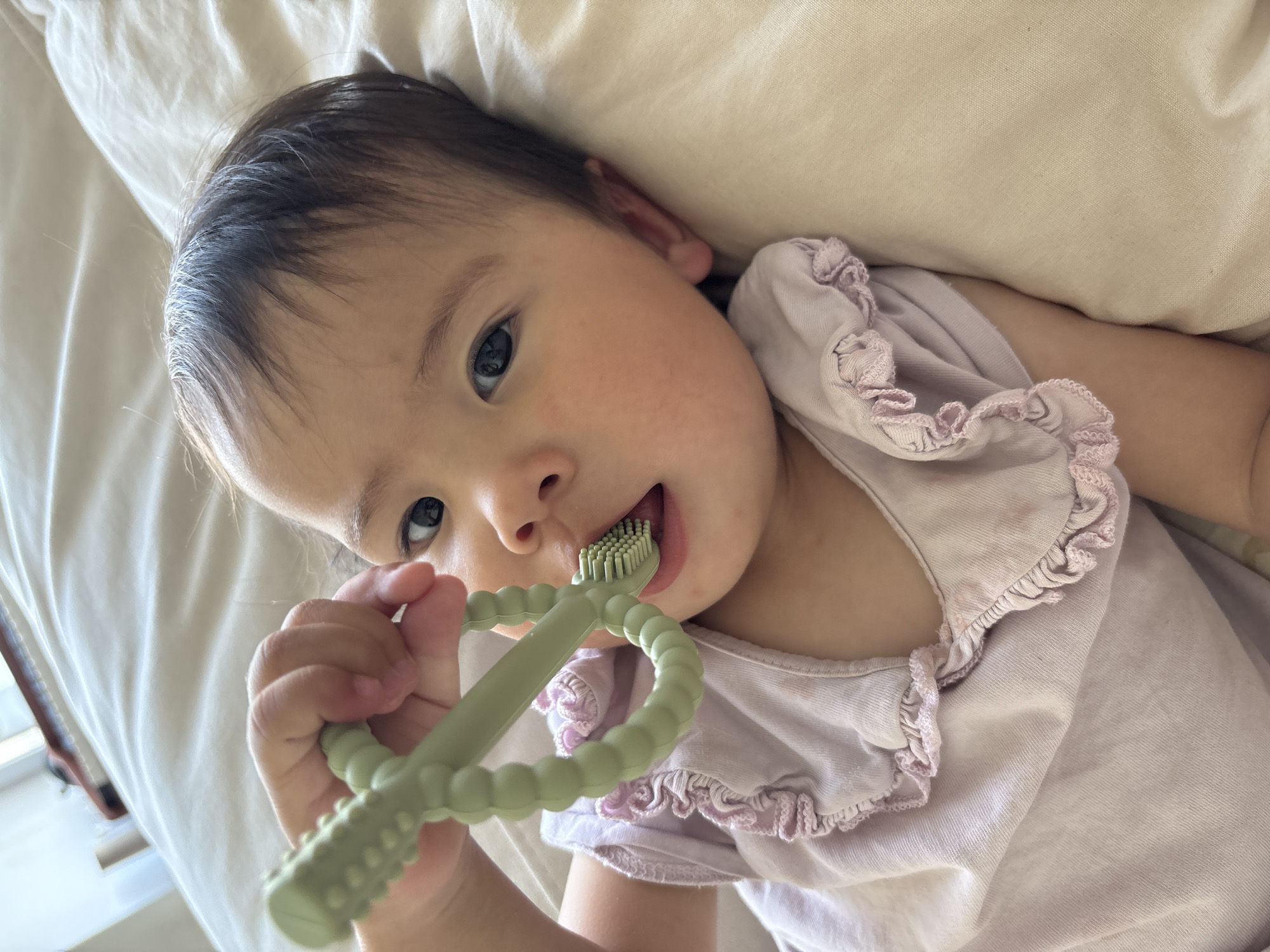 A baby is lying on a light-colored bed, possibly a pillow, holding a green toothbrush teether in their right hand and putting the brush part into their mouth. The baby is wearing a light pink shirt with a ruffled neckline. The location is possibly in Ембаево, Russia.