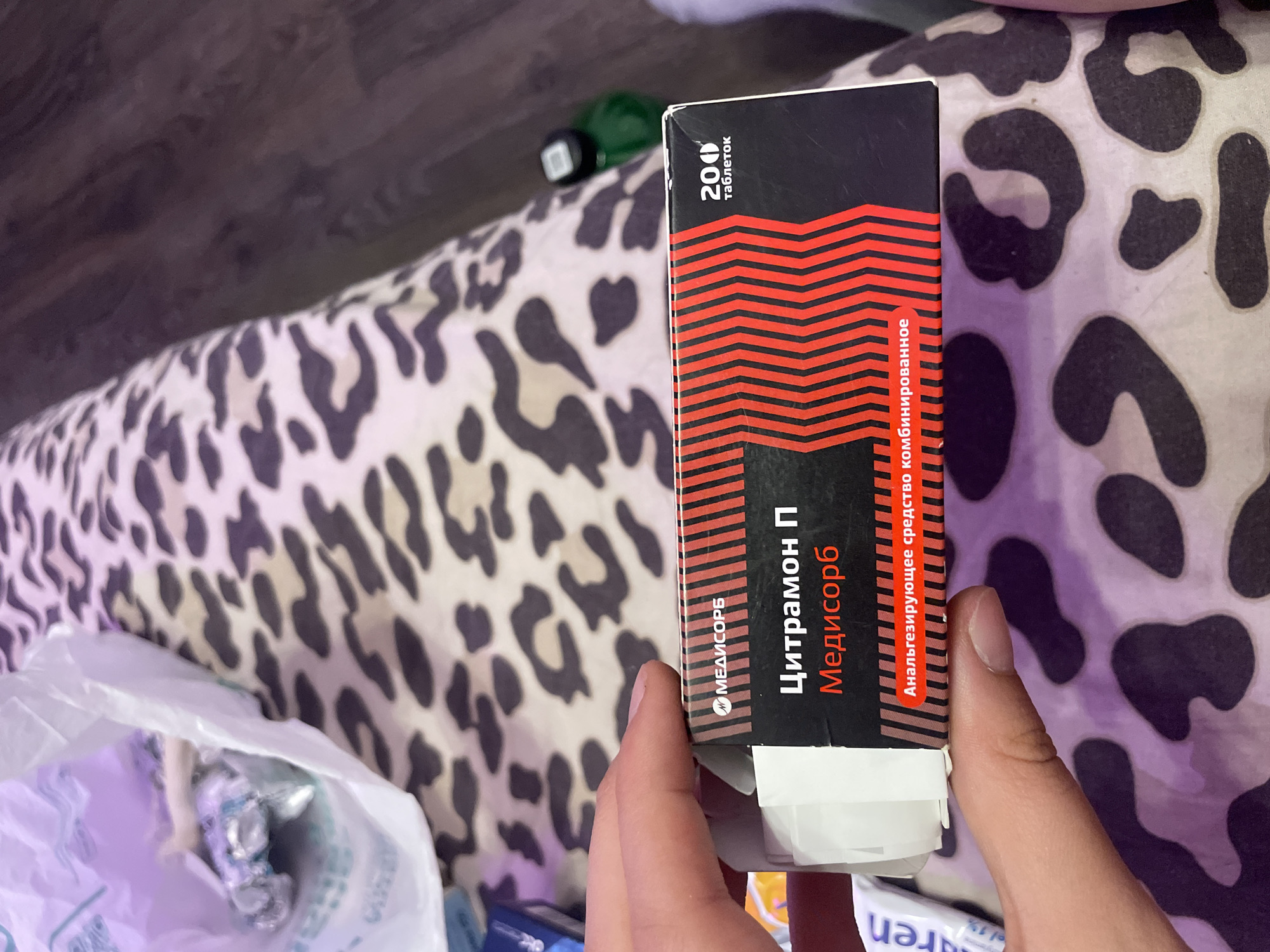 A person's hand is holding a box of "Citramon P" medication manufactured by "Medisorb". The box is predominantly black with red diagonal lines and white text. The text "200 таблеток" meaning "200 tablets" is visible in the upper right corner. The background consists of a leopard-print fabric and a dark surface, possibly a floor or table. The location is stated to be Vaganovo, Russia.