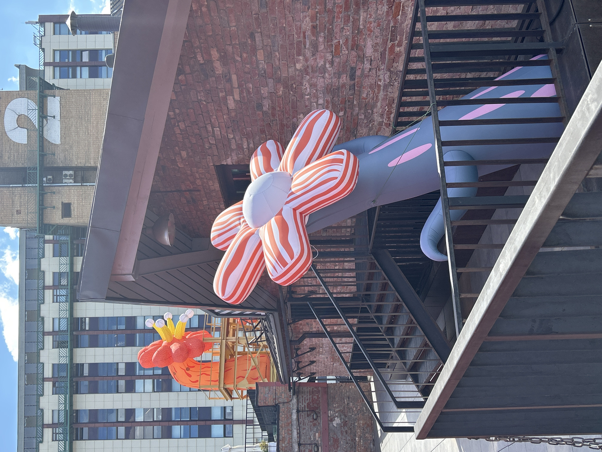 This is a medium-shot, eye-level, outdoor photograph taken on a bright, sunny day in an urban setting, possibly a rooftop. The foreground is dominated by a black metal railing that stretches across the bottom of the frame. Behind the railing, a set of dark metal stairs with black railing ascends.
The main subject of the photograph is a large, inflatable flower sculpture. The flower has petals with alternating orange and white stripes, and a white center. It is attached to a thick, gray stem adorned with irregular pink splotches and vertical dark pink stripes. The stem appears to be emerging from behind the stairs.
In the background, a red brick building forms the middle ground, with a dark brown pitched roof visible. Further in the background, two taller buildings are visible. The building to the left has a facade with alternating bands of white and dark windows, topped by a green railing. The building to the right is primarily made of brick and features a large, white, stylized number "2" on its upper section. Another taller building with dark windows and a white facade is partially visible behind it.
The sky above is blue with scattered white clouds. No people are visible in the image. The overall impression is one of an art installation in an urban environment.