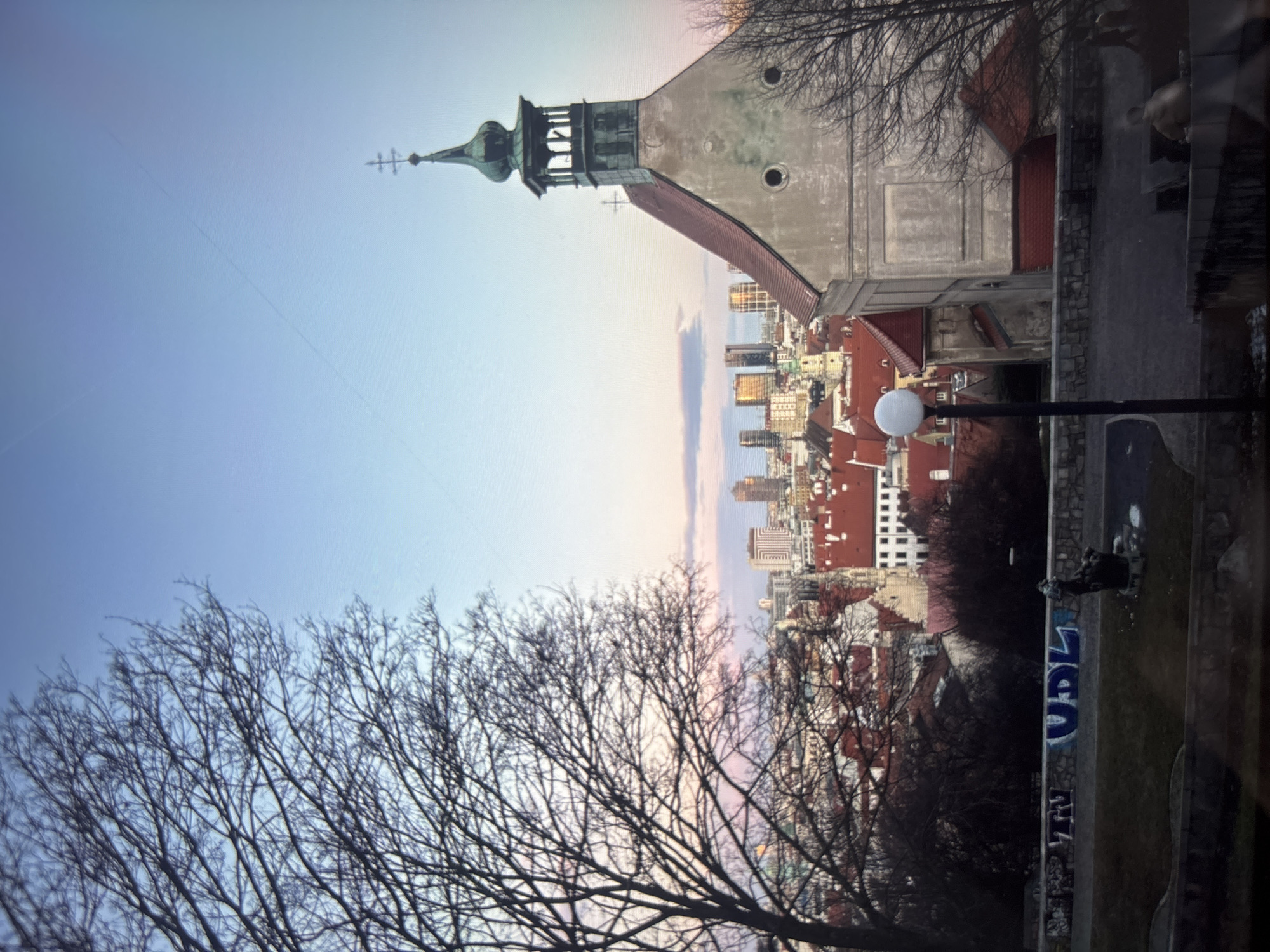 The image shows a cityscape with a view dominated by a church tower on the right, featuring a green spire and a light grey exterior. Adjacent to the church, a cityscape extends into the distance with numerous buildings, many with red-tiled roofs and taller skyscrapers in the background. In the foreground on the left, a large tree with bare branches partially obscures the cityscape. The sky transitions from a light blue to a pinkish hue towards the horizon. On the right, there's a lamp post and a graffiti-covered wall bordering a grassy area with a dark figure that is partially obscured. The location is Unknown, Unknown.