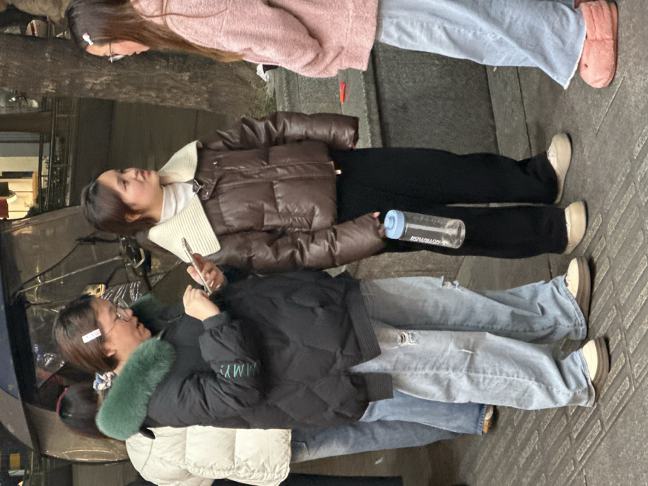 The image depicts an urban sidewalk in Jiangjin, China, featuring four individuals in casual winter clothing. From left to right, a partially visible person wears a white puffer jacket and light blue jeans. Adjacent to them, a woman with short dark hair, glasses, and a light-colored hair clip wears a black quilted puffer jacket with a green fur collar and light blue ripped jeans, operating a white smartphone with both hands. To her right, a second woman with dark hair wears a brown puffer jacket with a ribbed cream collar over a white turtleneck and black pants; she holds a clear plastic water bottle with a blue lid in her right hand and directs her gaze toward the rightmost individual. On the far right, a third woman with long dark hair and glasses, wearing a pink fuzzy jacket, light blue wide-leg jeans, and pink fluffy slippers, faces the second woman, suggesting an interaction. The immediate environment consists of a paved sidewalk with visible tactile strips. In the background, obscured by the individuals, a dark three-wheeled vehicle with a transparent plastic cover and a tree trunk are discernible. Distant blurred building facades with internal illumination are also present.