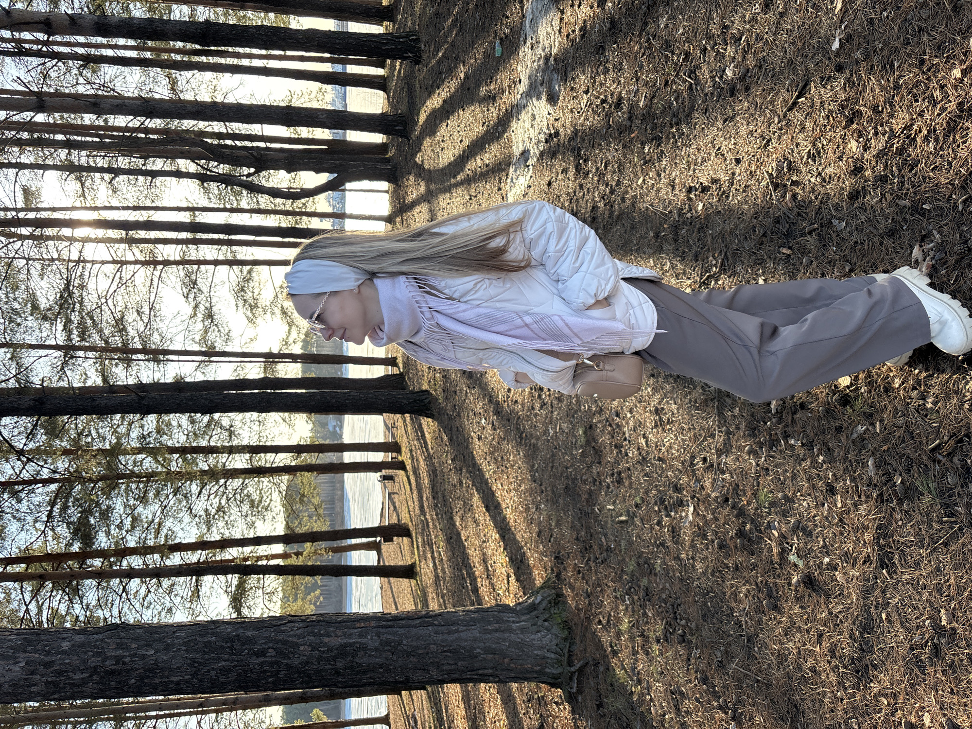 The image shows a woman standing in a wooded area, possibly a park. In the background, there's a glimpse of water, suggesting a lake or river. The woman is wearing a white puffer jacket, brown pants, and white shoes. She has long blonde hair and is wearing a white headband and glasses. The ground is covered with pine needles and the shadows of trees. The light suggests it is a sunny day. The trees are tall and thin. Given the mention of the city of Chaikovsky, Russia, it is plausible that the location is a park near the Kama River or another body of water in the area.