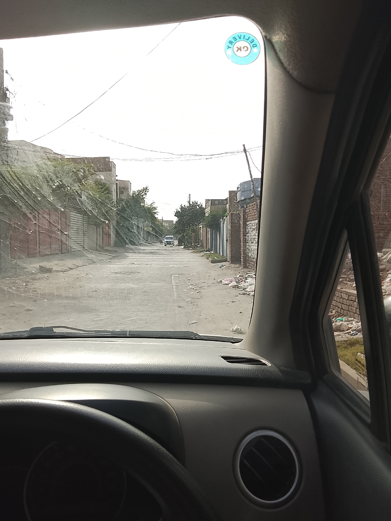 The image shows a view from inside a car, looking out onto a street in Gulgasht Colony, Pakistan. The street is lined with buildings, some with metal gates or fences. Litter is visible along the sides of the road. A car is seen in the distance. The car's dashboard and steering wheel are partially visible. A round air vent is seen. A sticker is located at the top of the front window. The sky is overcast.