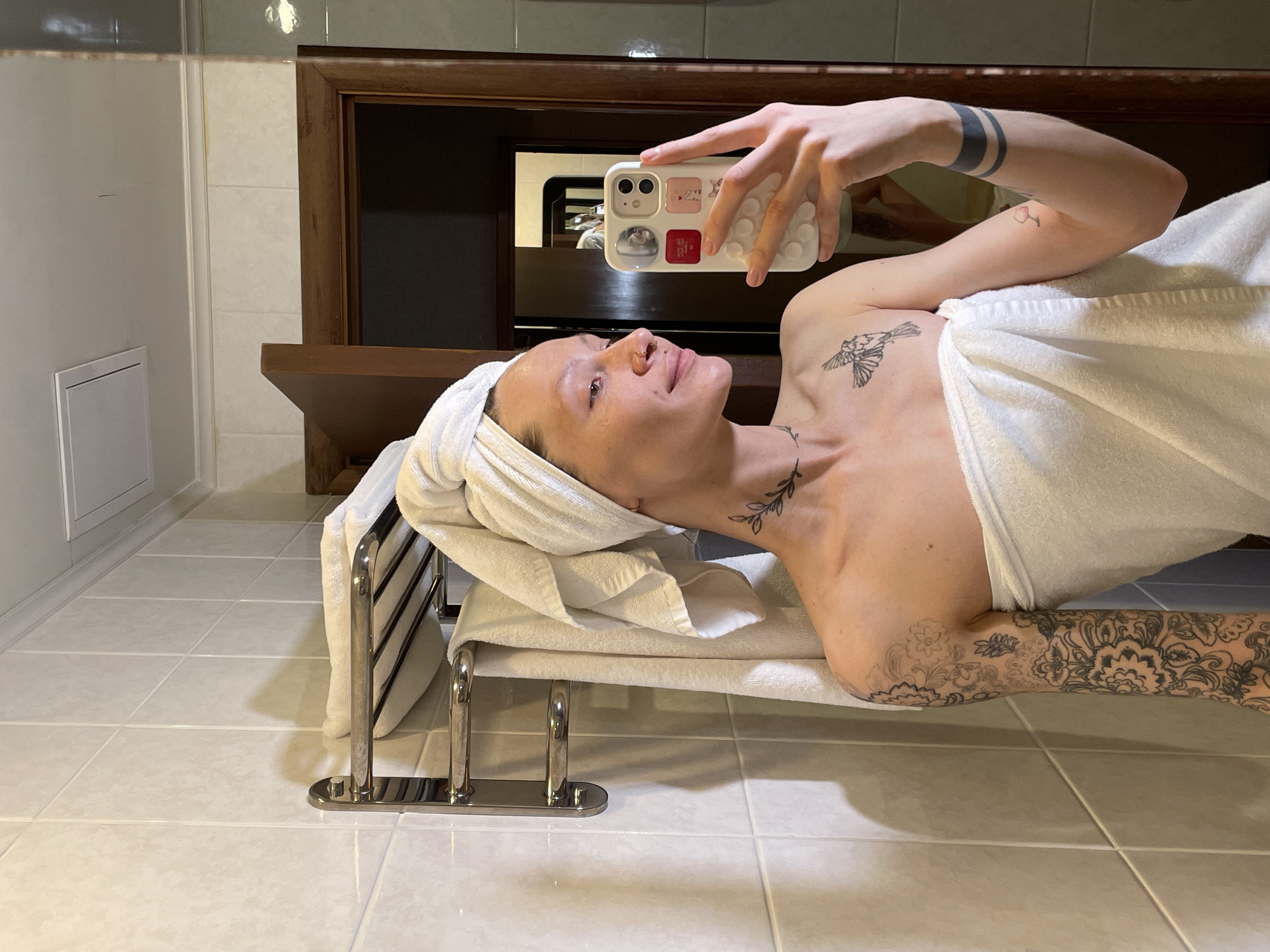 A person is lying on a white towel on a silver rack. The person is partially covered by the towel, and they have a white towel wrapped around their head. The person is holding a cell phone in their right hand and taking a selfie. The person has several tattoos, including one of a bird on their chest, one of a vine on their neck, and extensive tattoos covering their left arm. The scene is set in a bathroom, with light-colored tile flooring and walls. There is a dark brown wooden cabinet or shelf unit mounted on the wall behind the person. The exact location in Naberezhnye Chelny, Russia, cannot be determined from the image alone.