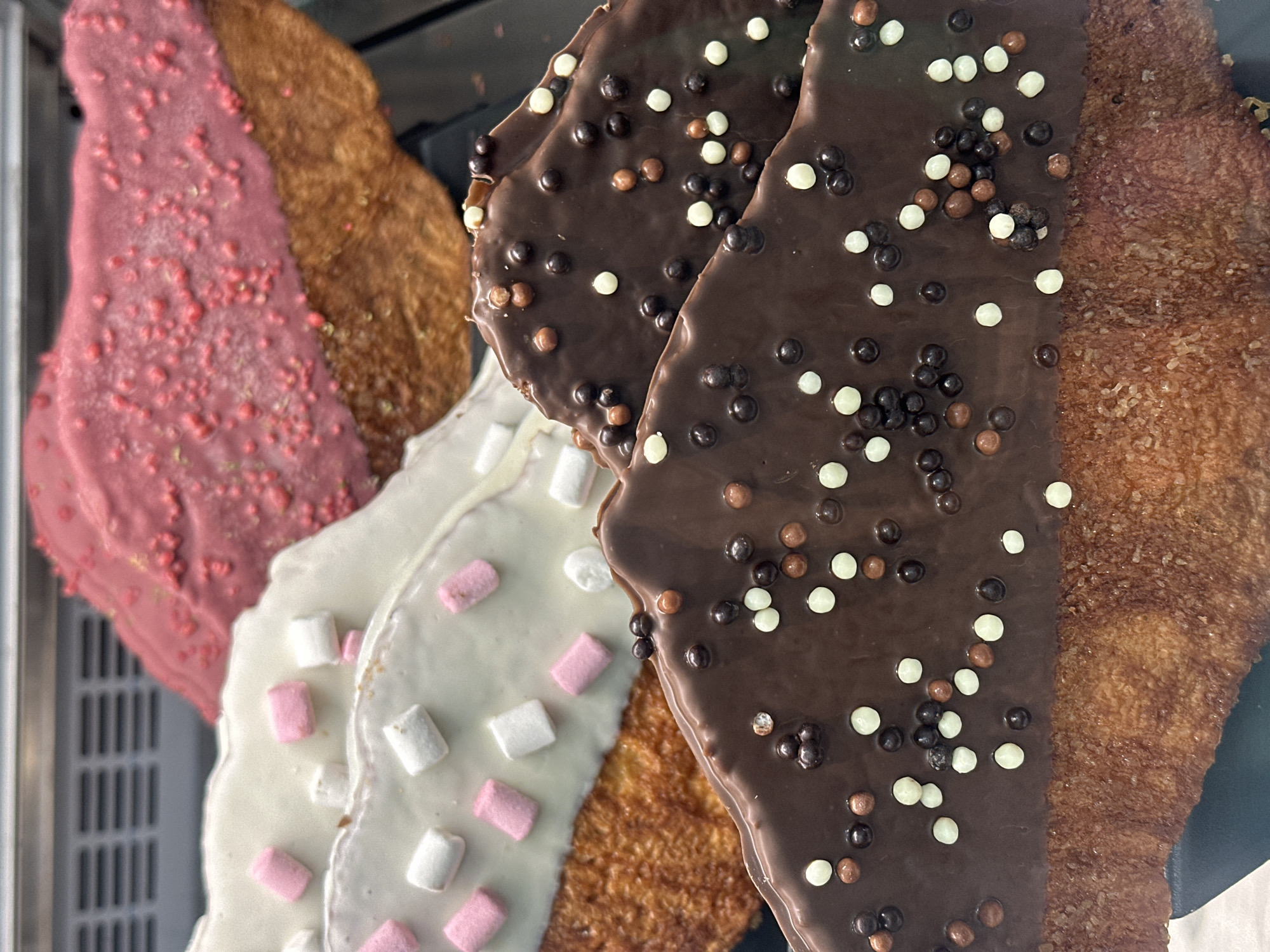 The image shows a close-up view of several churros variations on display, likely in a pastry shop or cafe. Each churro has a different topping or coating on one half, including pink with sprinkles, white with mini marshmallows, and dark chocolate with colored sprinkles. The uncoated halves of the churros have a light-brown, fried texture. The scene is likely in Madrid, Spain, given churros are a popular treat in the city.